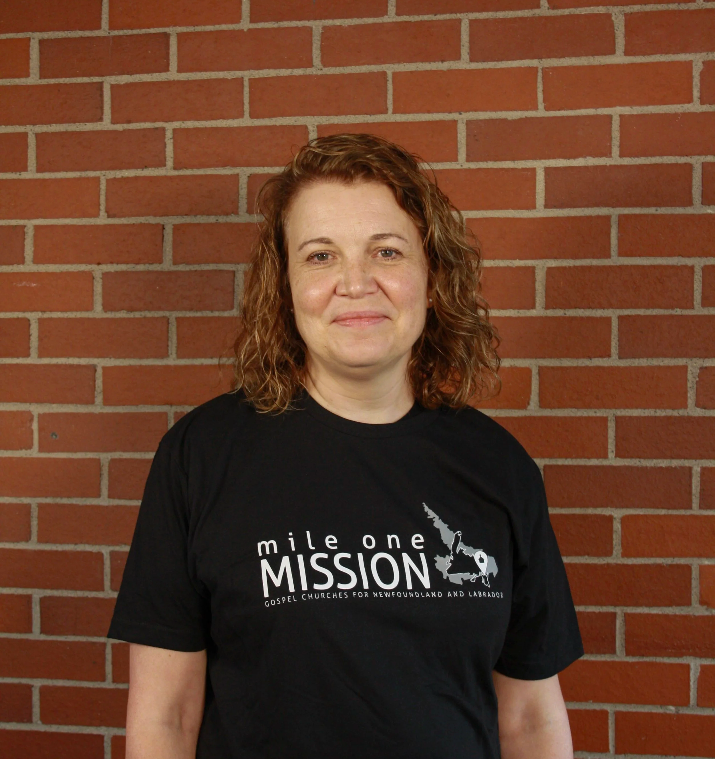 Woman with red hair standing in front of a brick wall wearing a black shirt with white text promoting gospel churches in Newfoundland and Labrador.