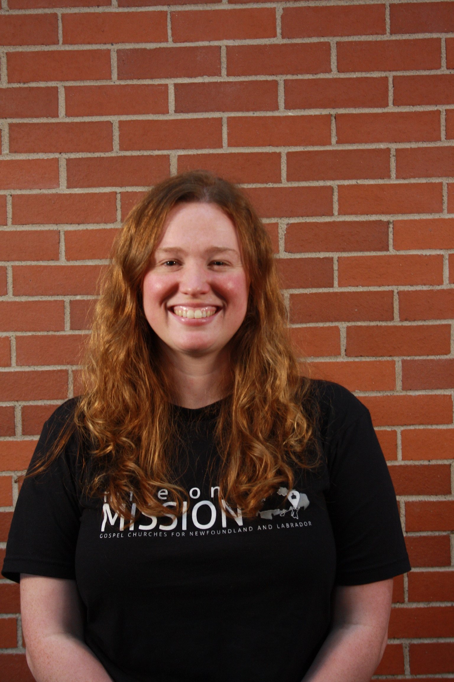 A smiling woman with long wavy red hair is standing in front of a red brick wall. She is wearing a black T-shirt with white text and a graphic.
