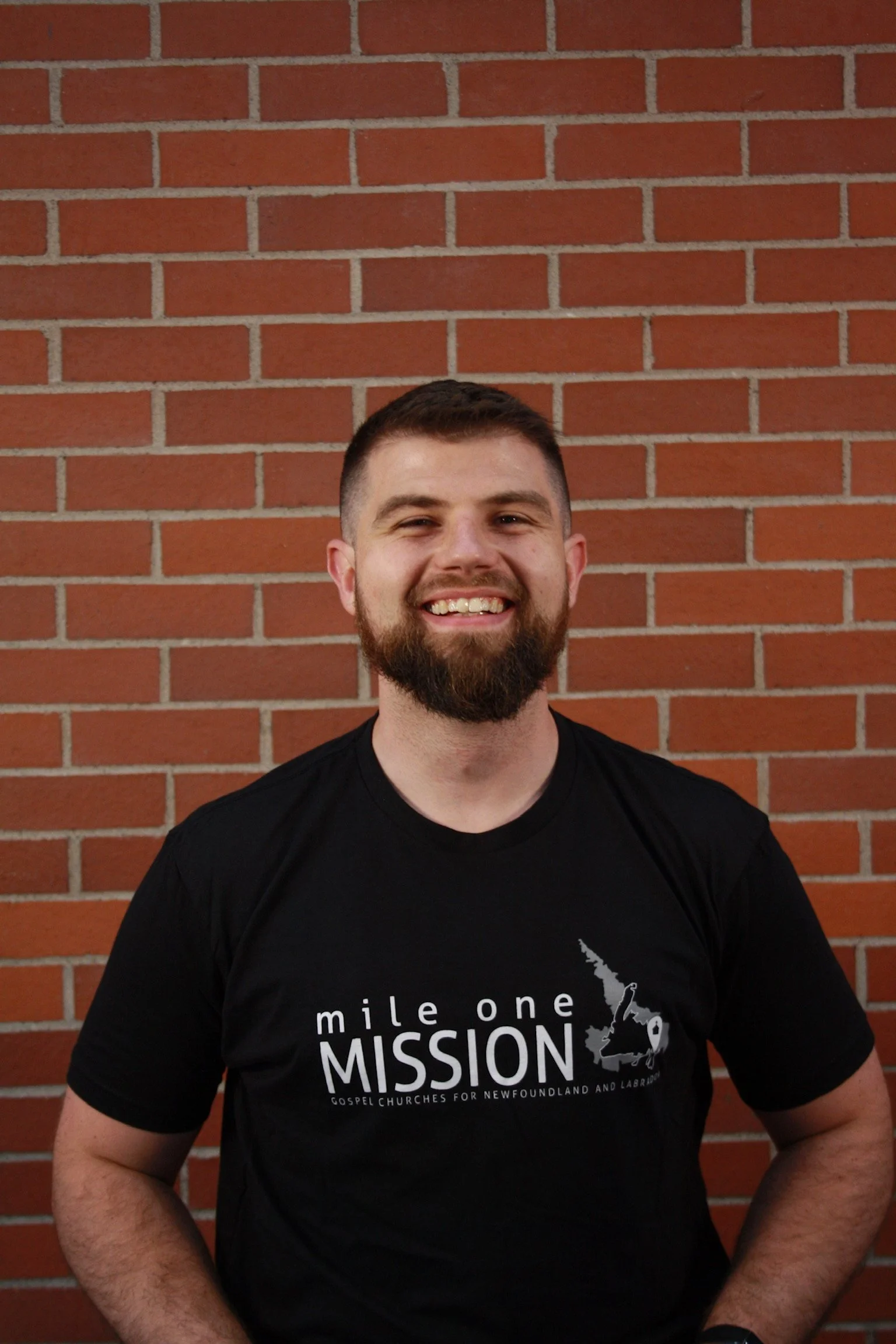 A smiling man with short brown hair and a beard, wearing a black T-shirt with a logo, standing in front of a red brick wall.