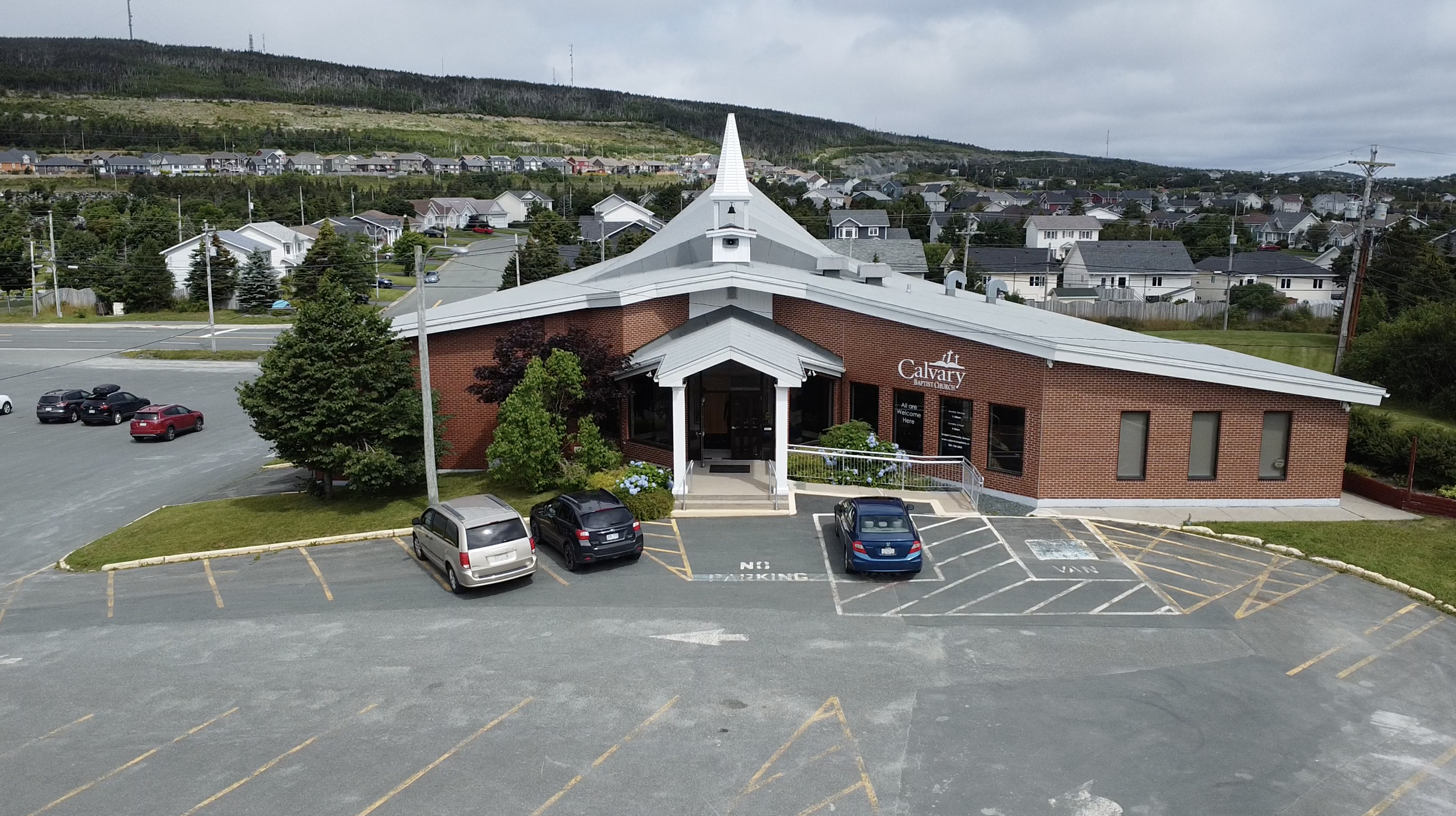 A brick church building with a white roof and steeple, parking lot with several cars, and a background of residential houses and green hillside.