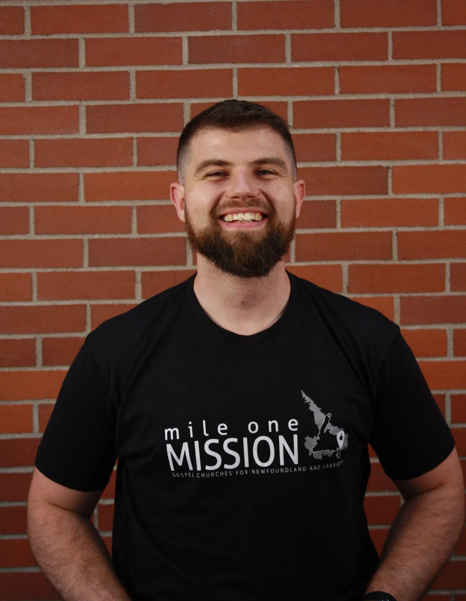 A man with short hair and a beard smiling in front of a brick wall, wearing a black T-shirt with white text that says "mile one MISSION" and a map outline of New Zealand with a church key.