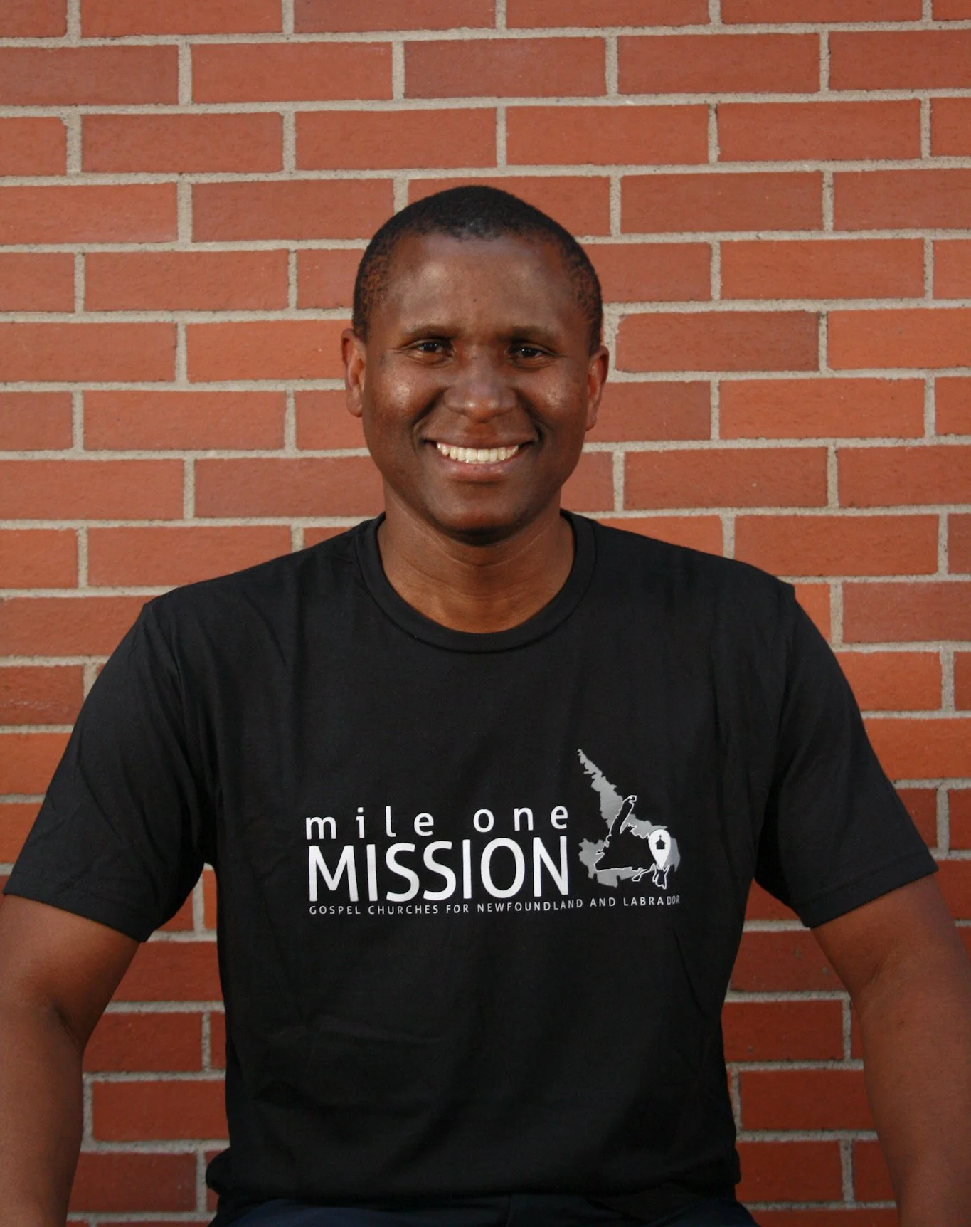 A smiling man sitting against a brick wall, wearing a black T-shirt with white text and graphics about a mission for gospel churches in Newfoundland and Labrador.