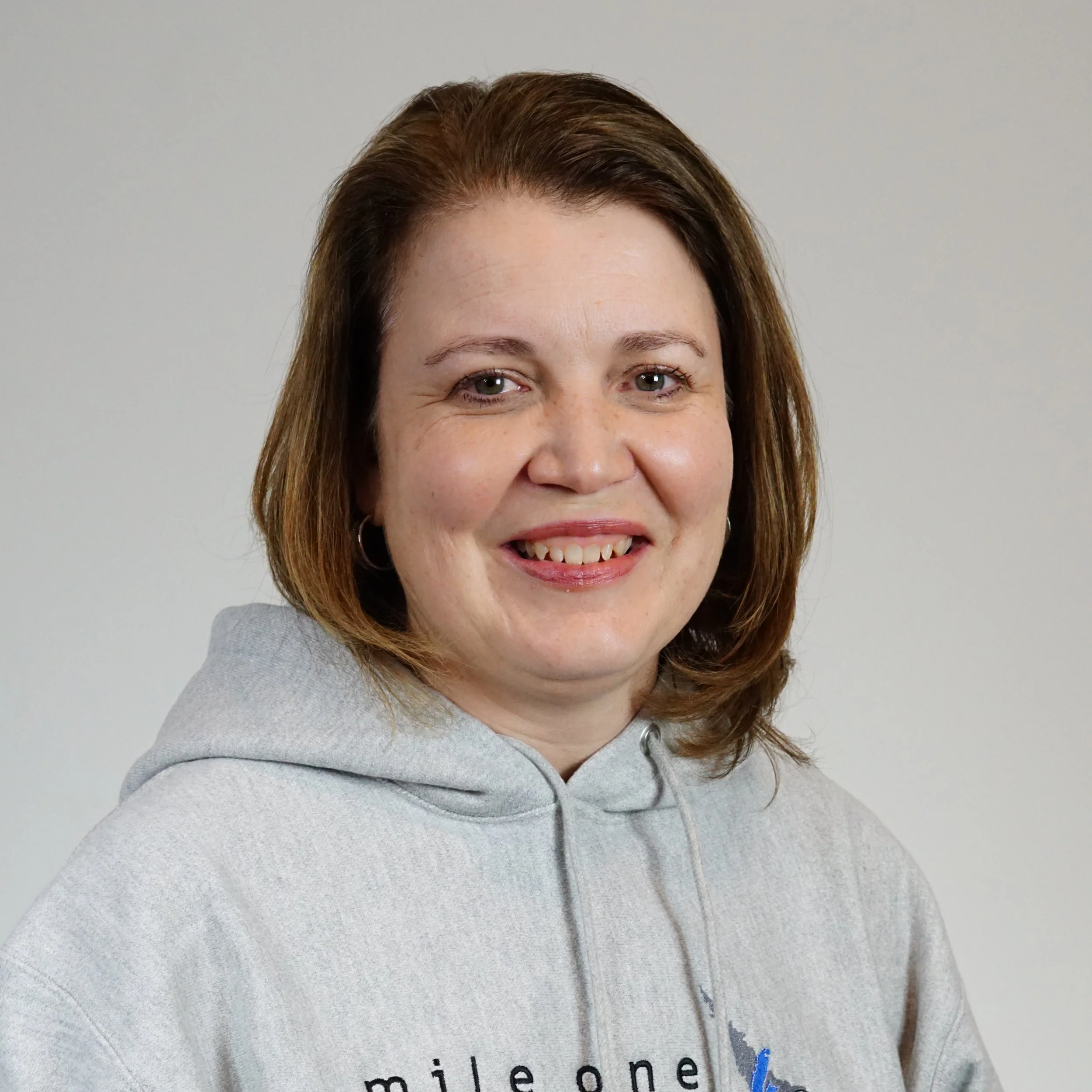 Close-up of a woman with shoulder-length brown hair, smiling, wearing a gray hoodie with text partially visible.