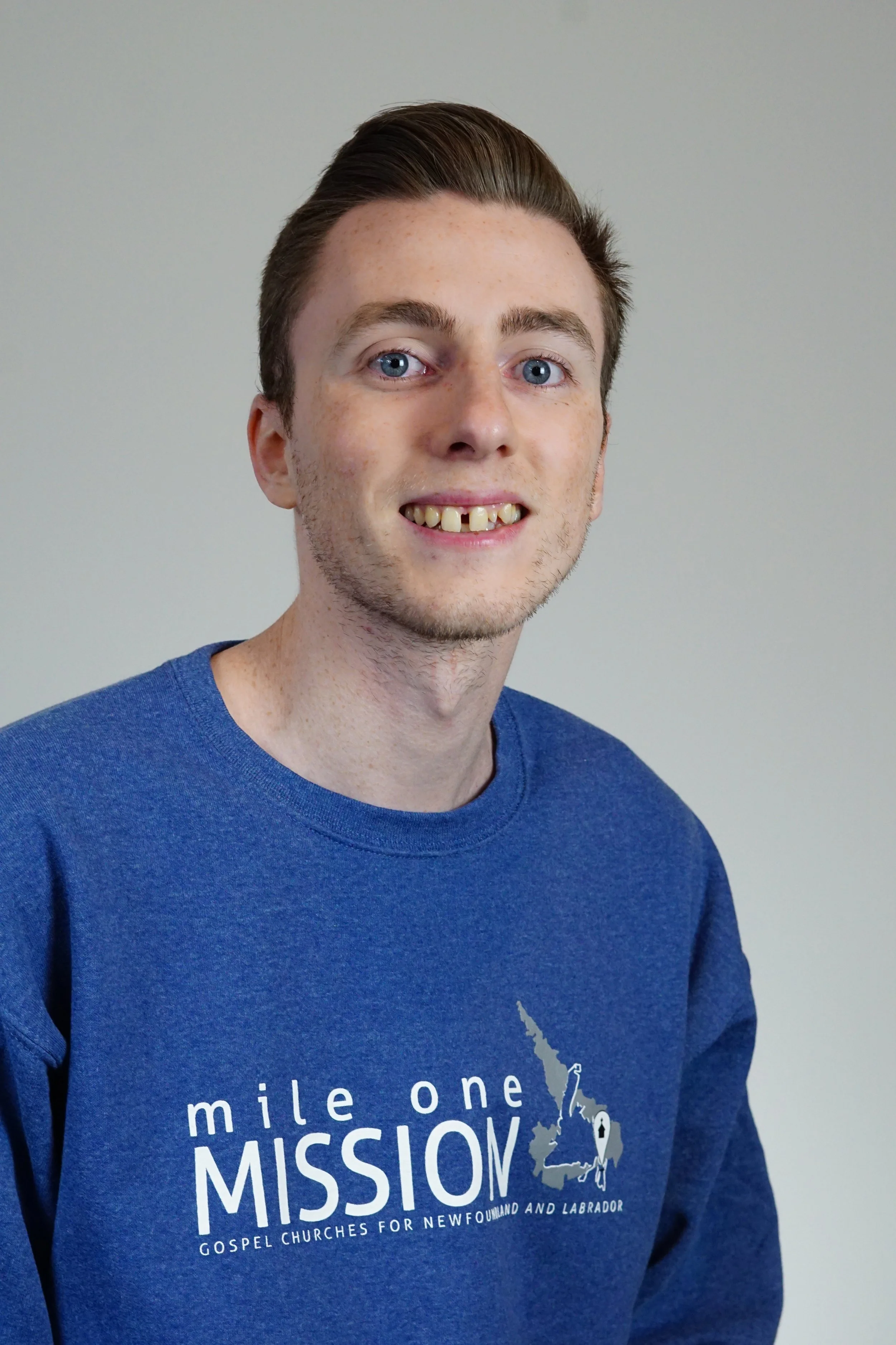 Young man wearing a blue sweatshirt with the words 'mile one mission' and a map of Newfoundland and Labrador printed on it, standing against a plain light-colored background.