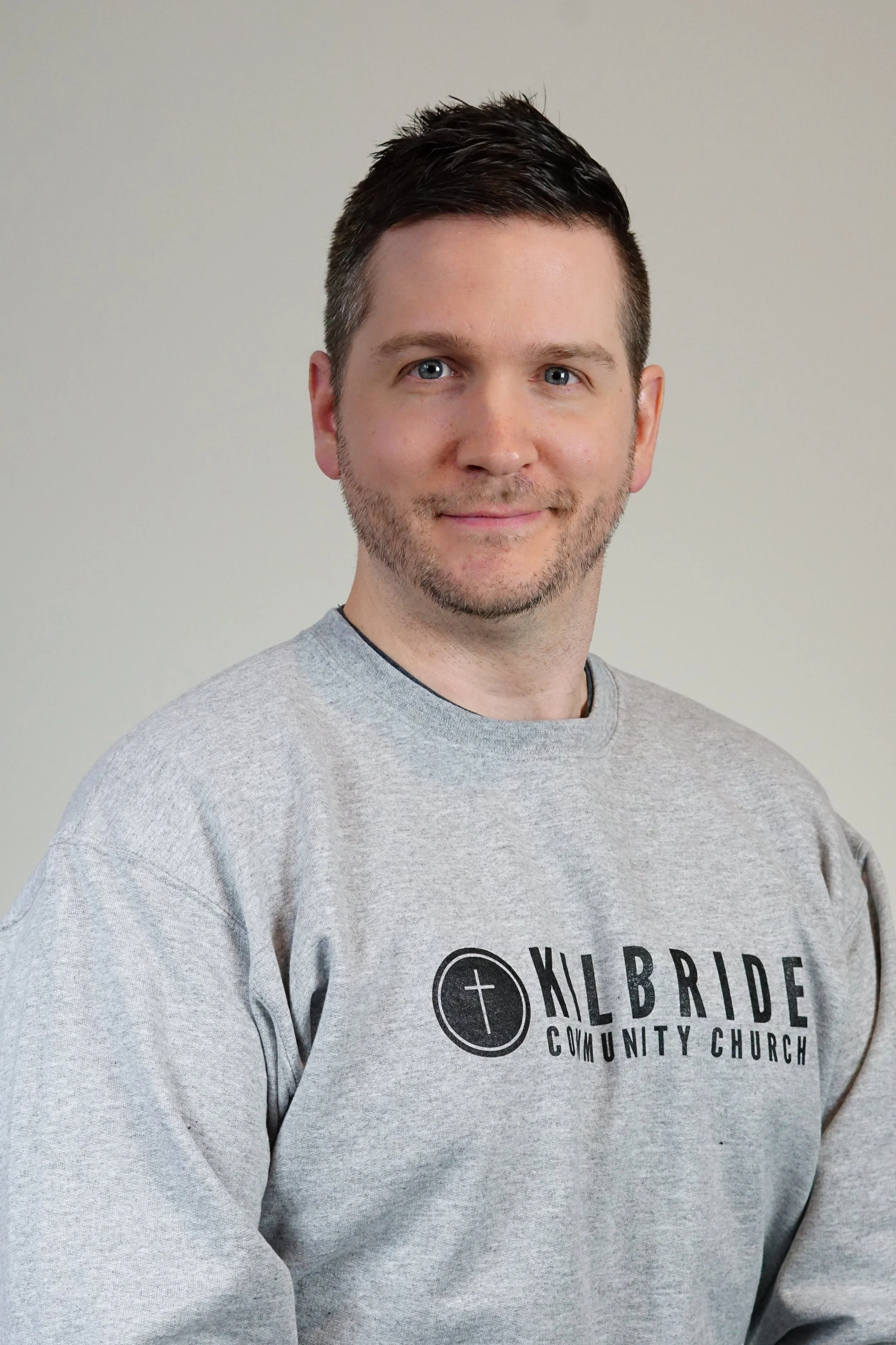 A man with short dark hair and a beard, wearing a gray sweatshirt with the logo and text of 'KL BIBLE COMMUNITY CHURCH', posing against a plain light gray background.