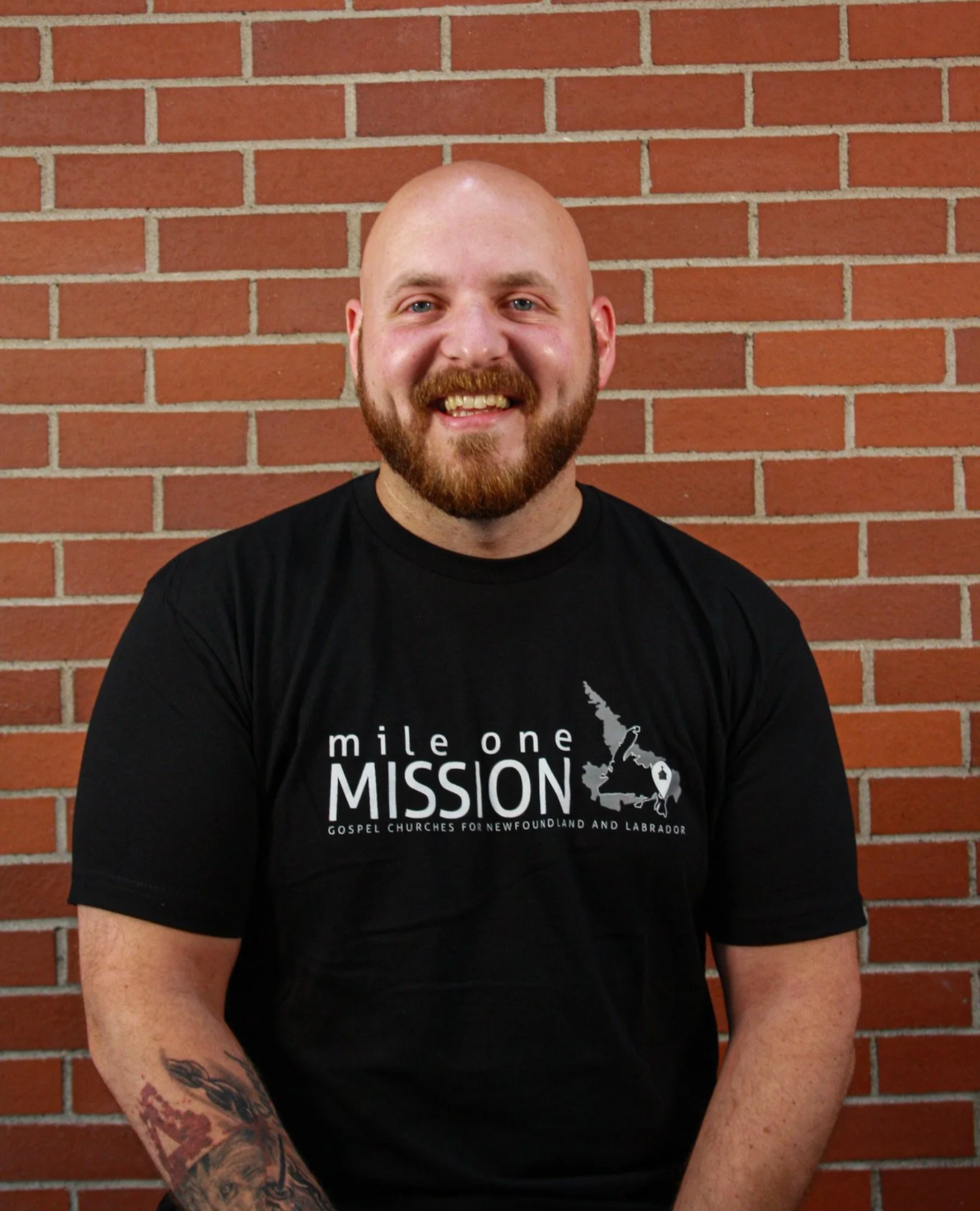 A smiling man with a beard and tattoos on his left arm, wearing a black t-shirt with a graphic and text, standing in front of a brick wall.