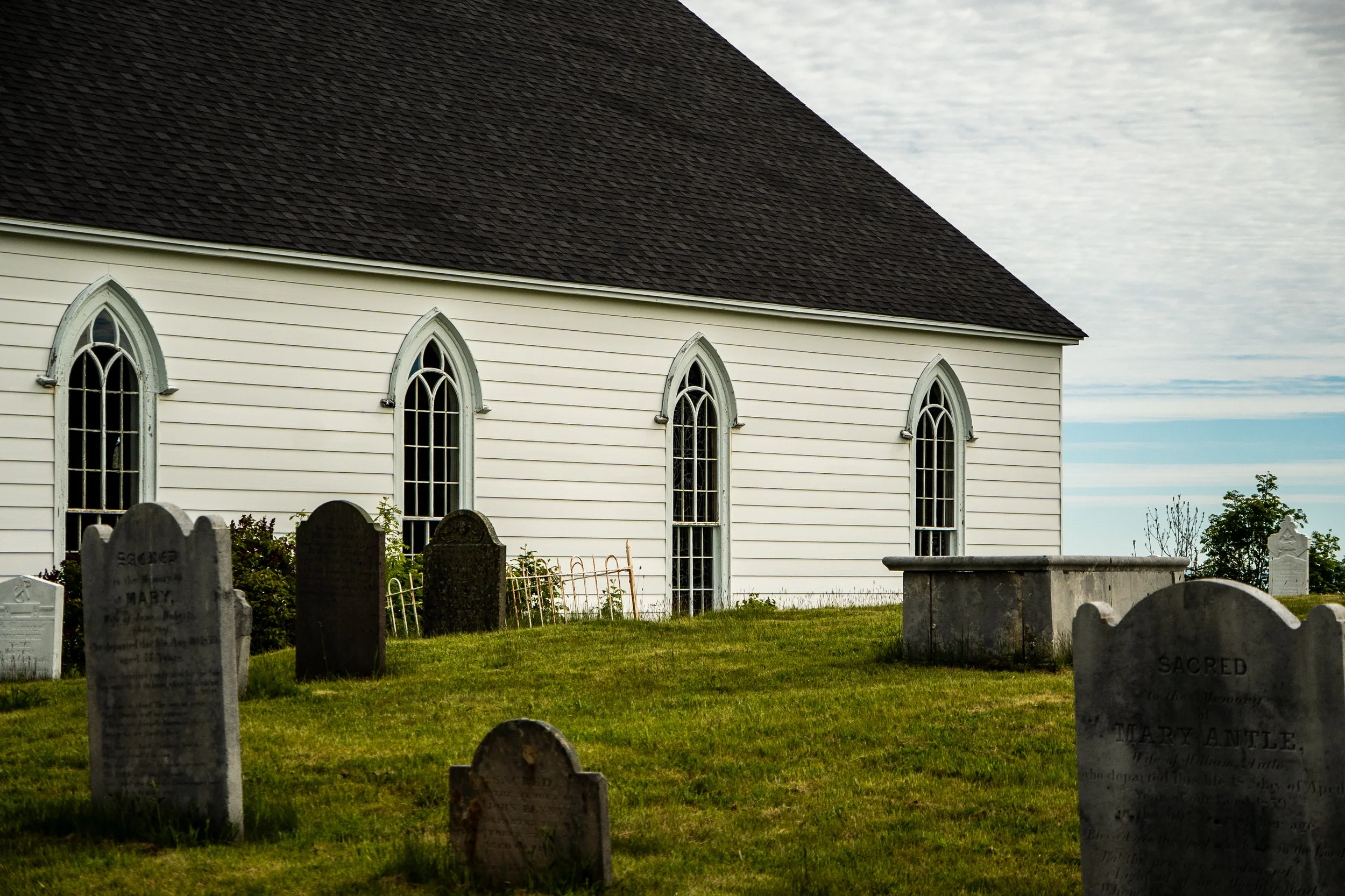 A white church with arched windows and a steep dark roof, surrounded by old tombstones in a graveyard, with partly cloudy sky in the background.