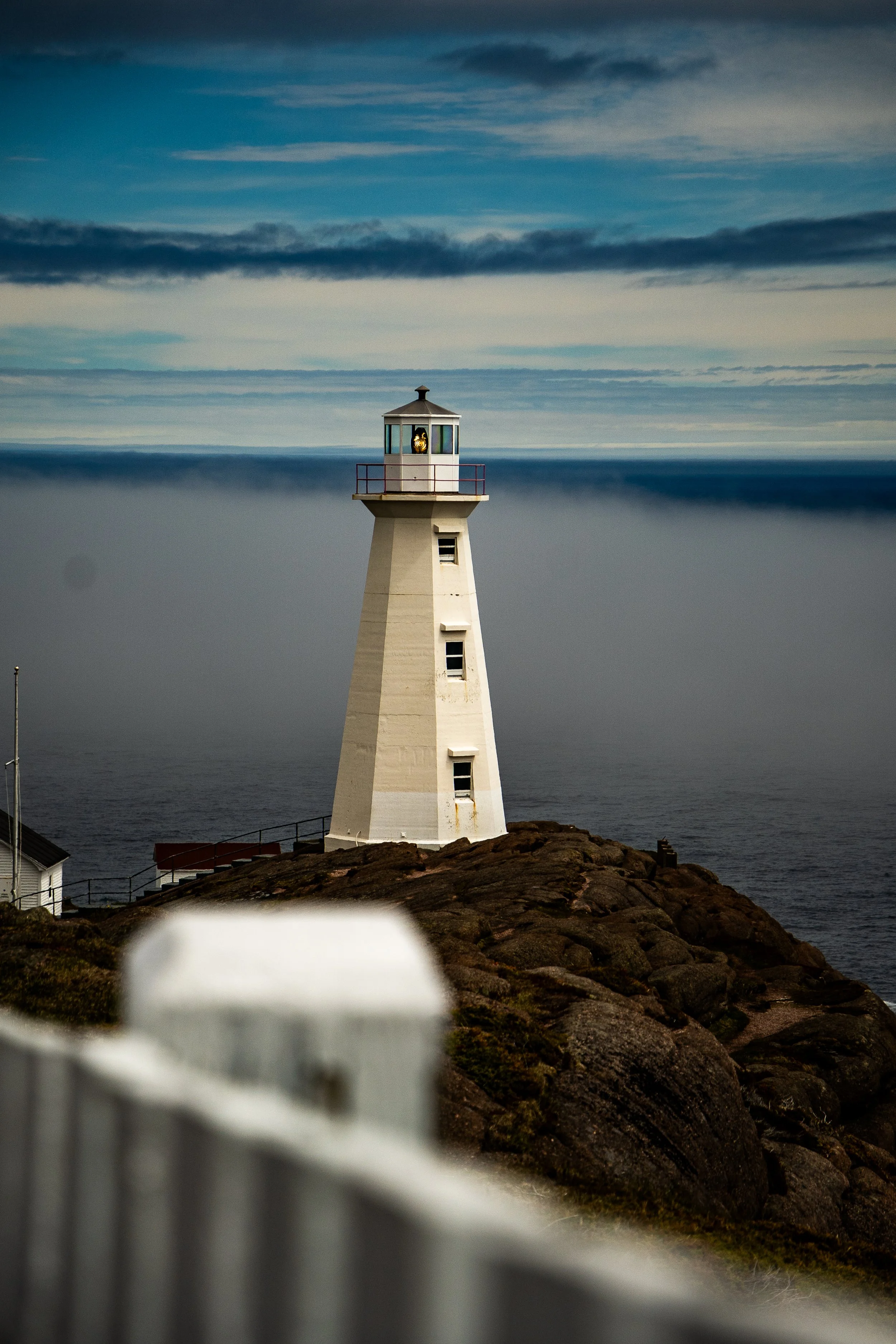 A lighthouse stands on a rocky cliff overlooking the ocean, with a partly cloudy sky overhead.