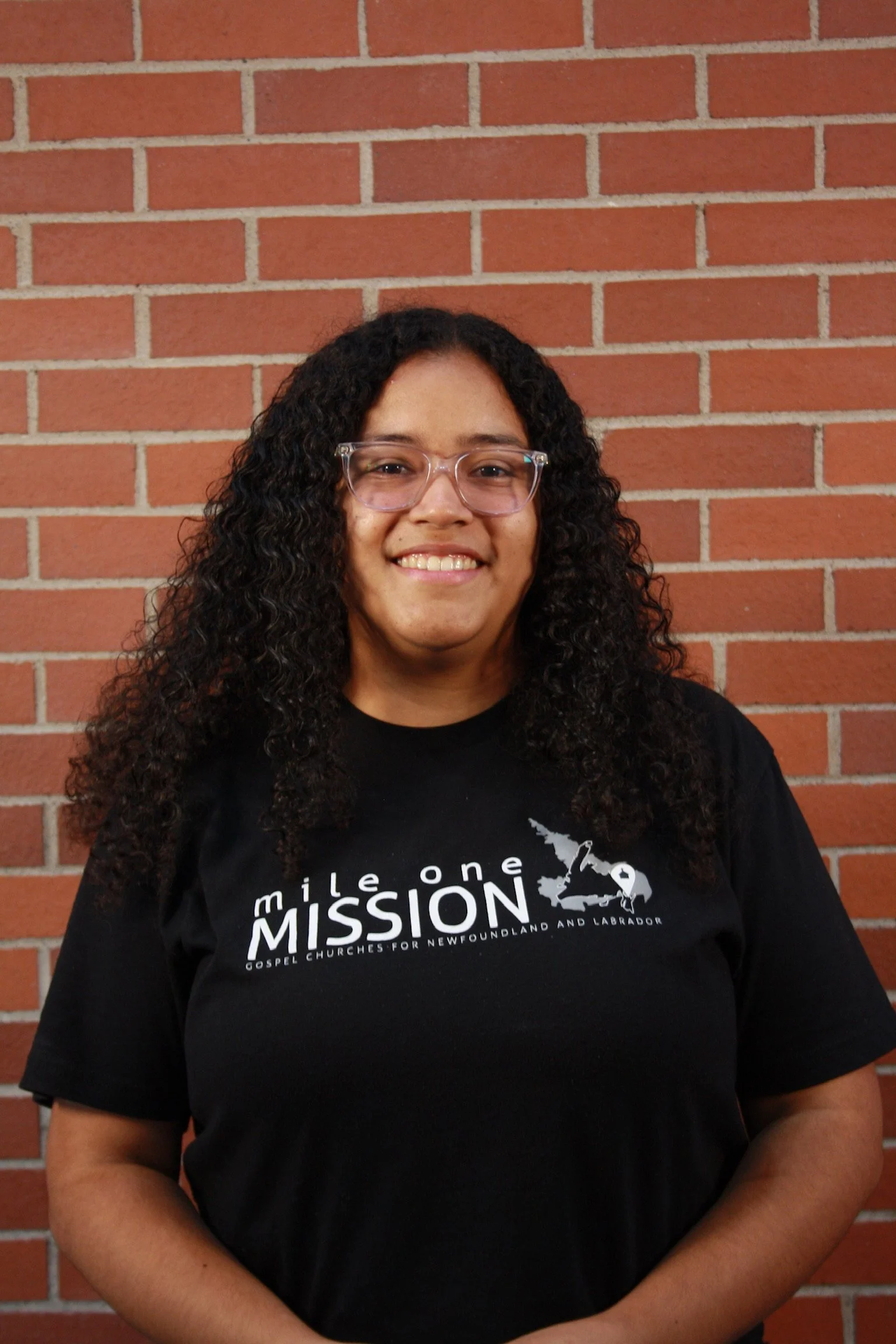 A woman with curly hair, glasses, and a black T-shirt standing in front of a brick wall.