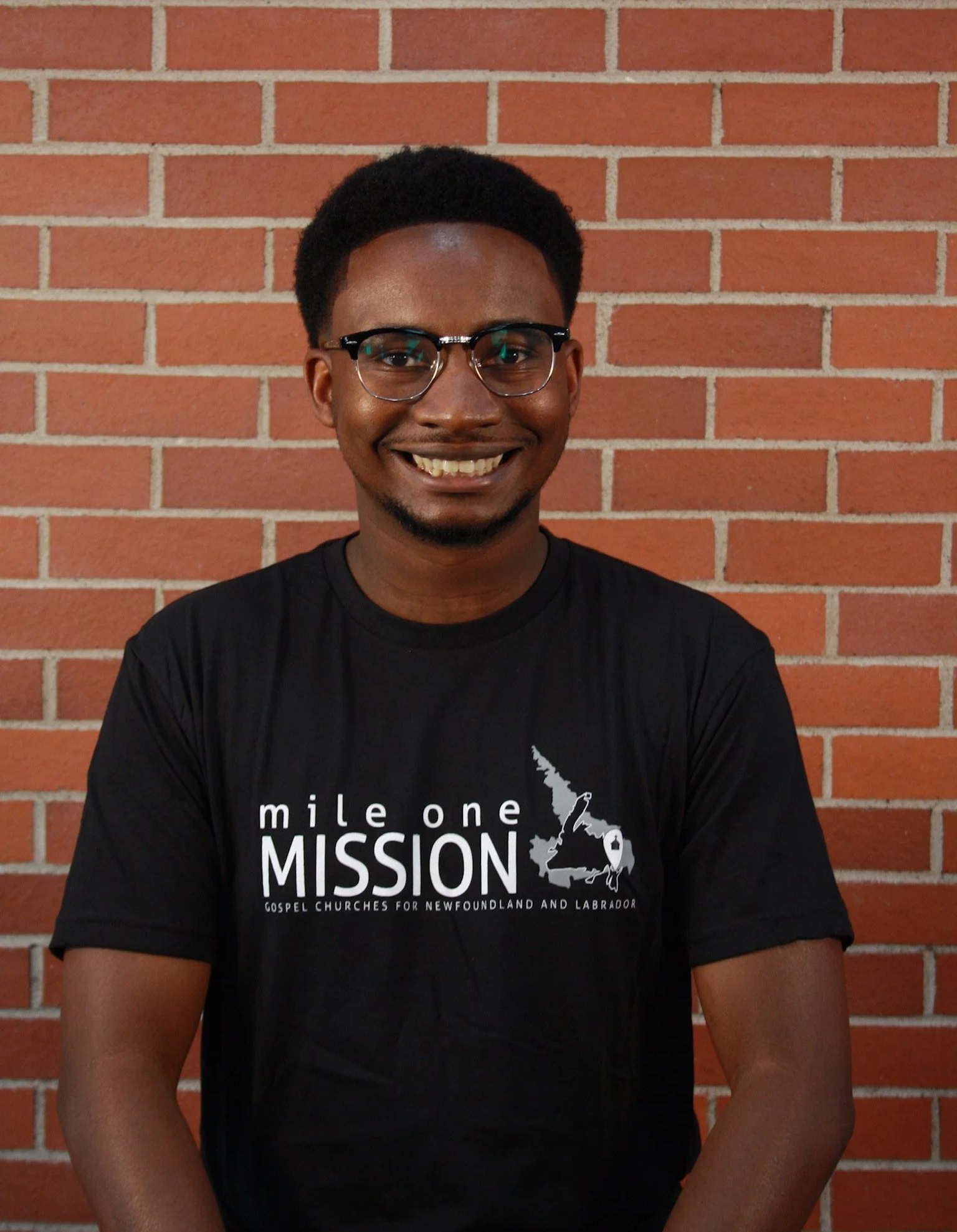 A smiling young man wearing glasses and a black t-shirt standing in front of a brick wall.