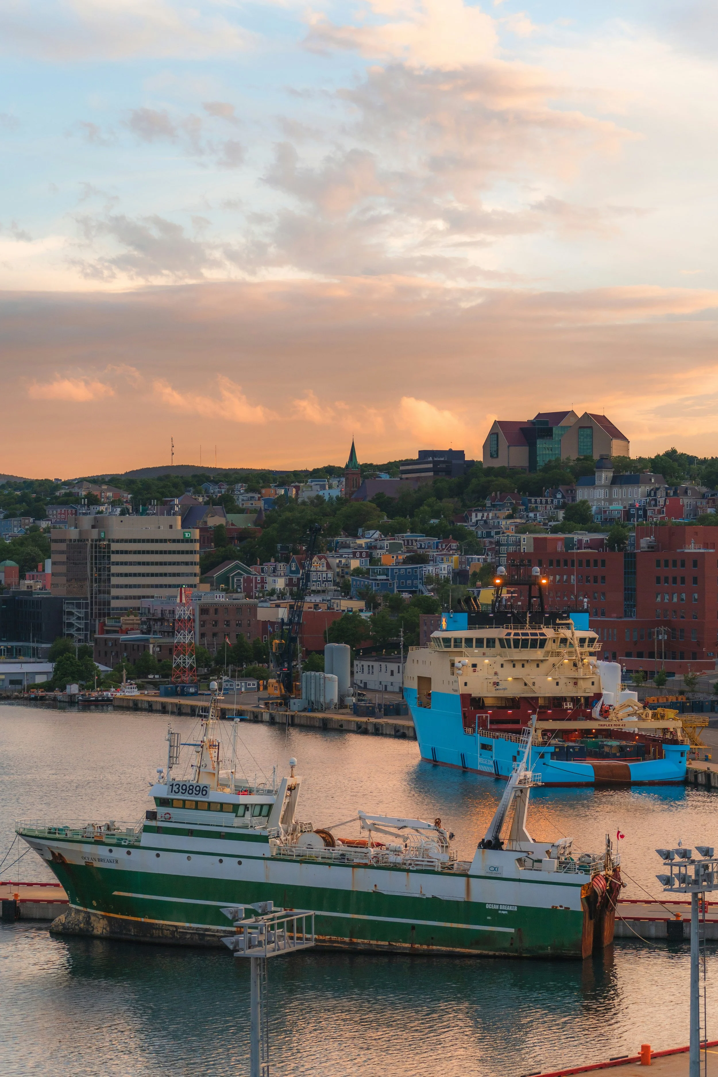 Harbor scene at sunset with boats docked, high-rise buildings, and a scenic hilltop with houses and a church steeple.