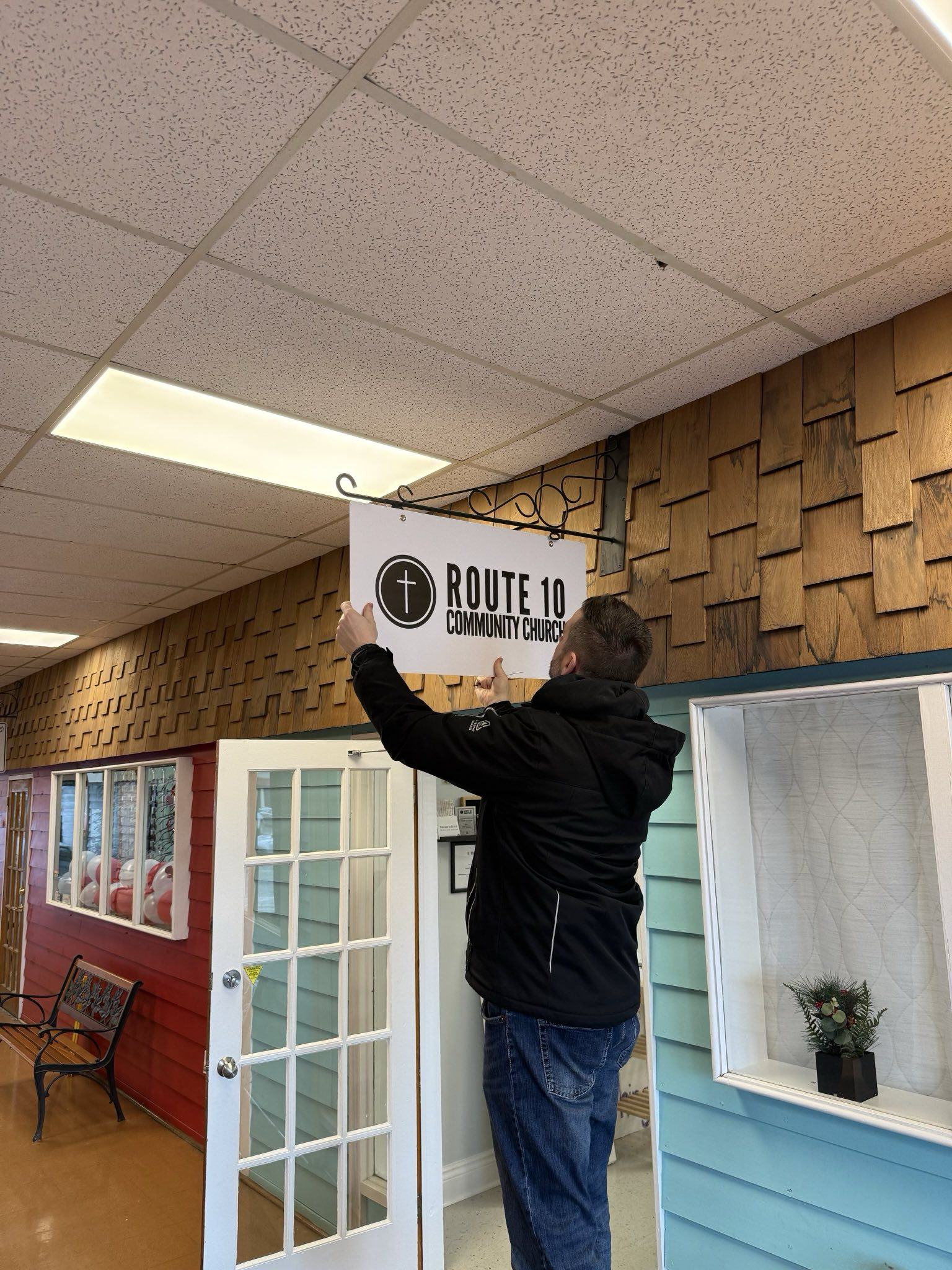 A man holding a sign that says 'Route 10 Community Church' inside a store or mall with decorated walls and furniture.