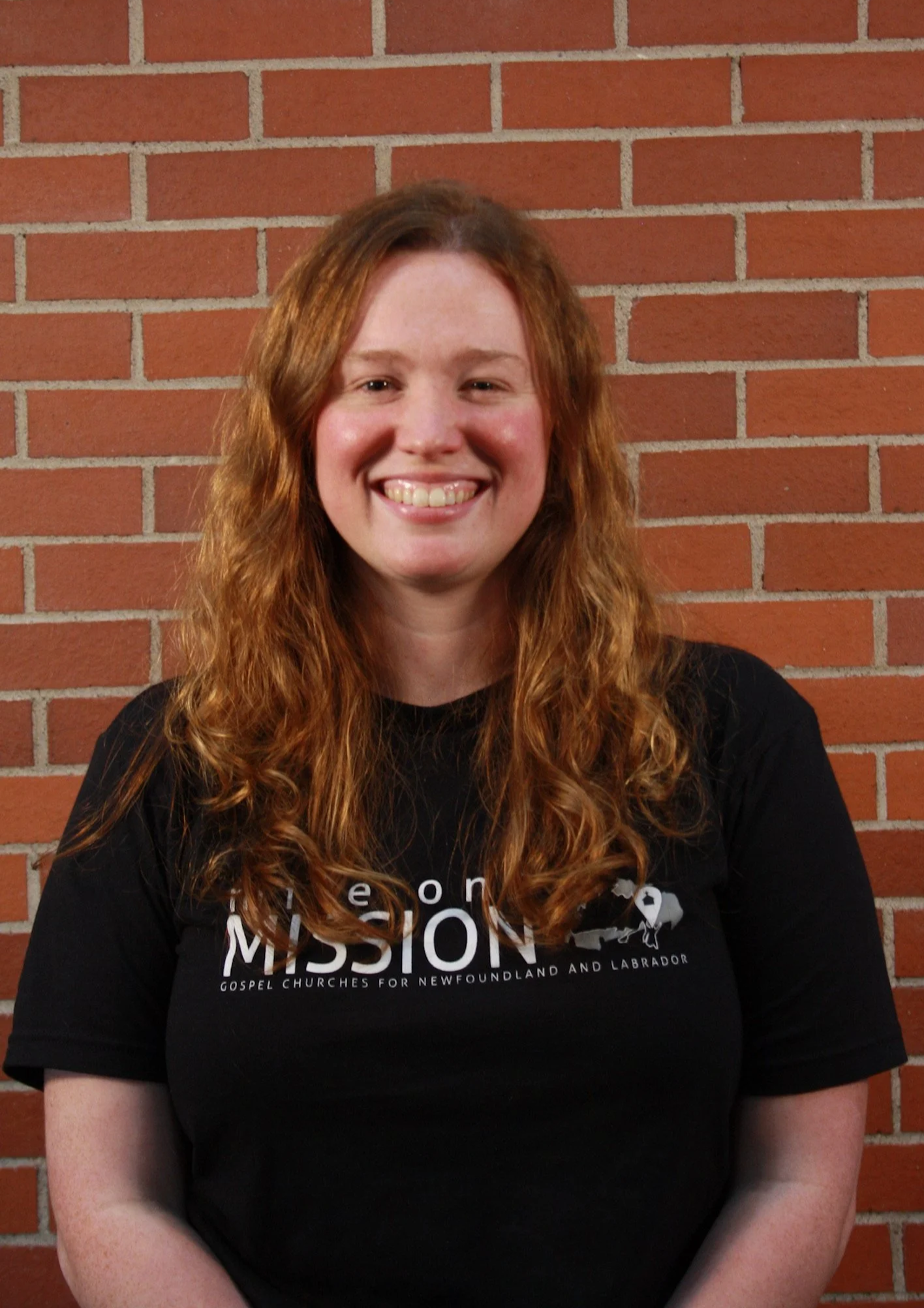 A woman with long curly red hair smiling while standing against a brick wall, wearing a black t-shirt with white text and graphics.