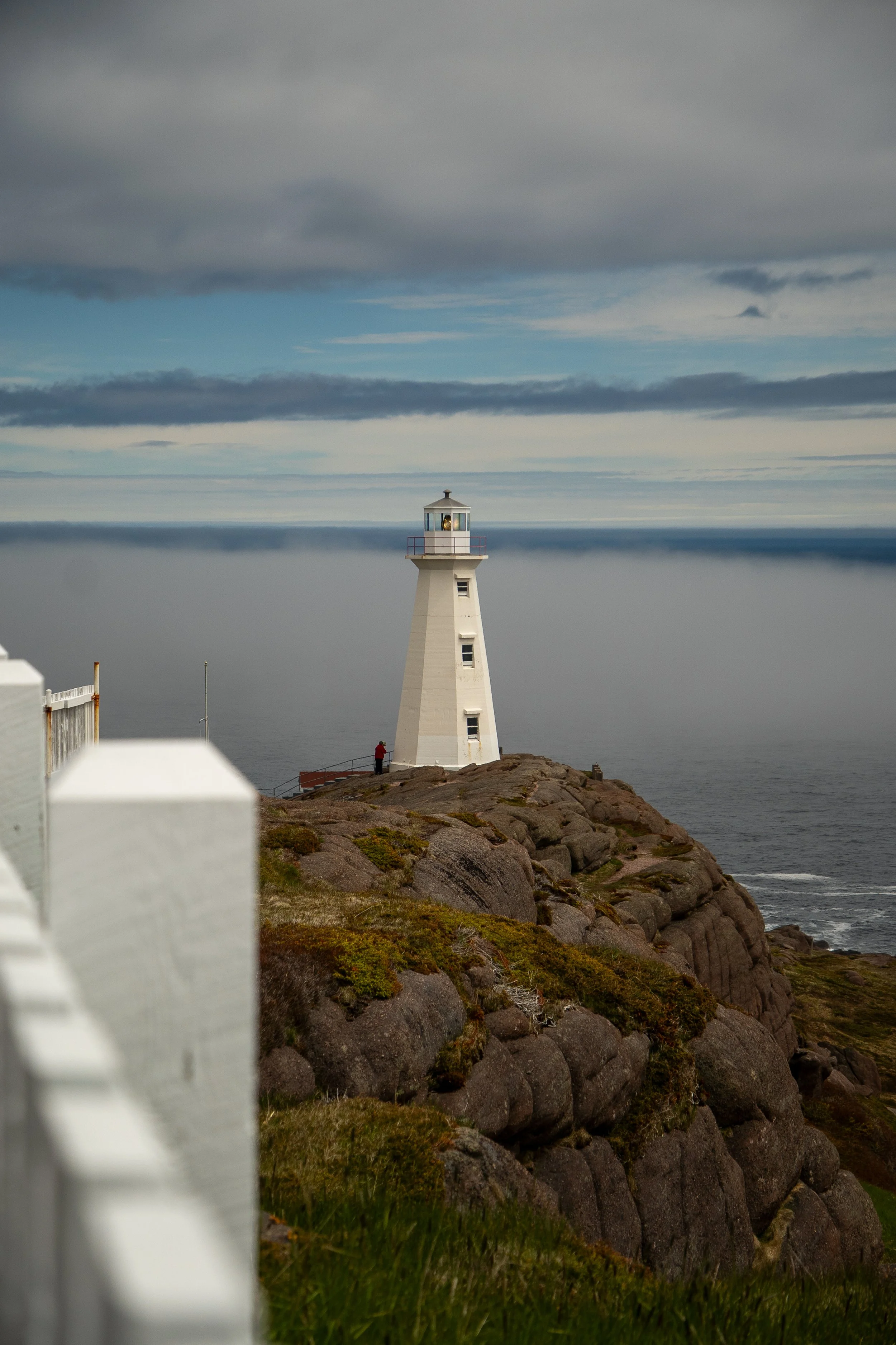 White lighthouse on rocky coast with cloudy sky and ocean in the background, partly obscured by a white fence in the foreground.