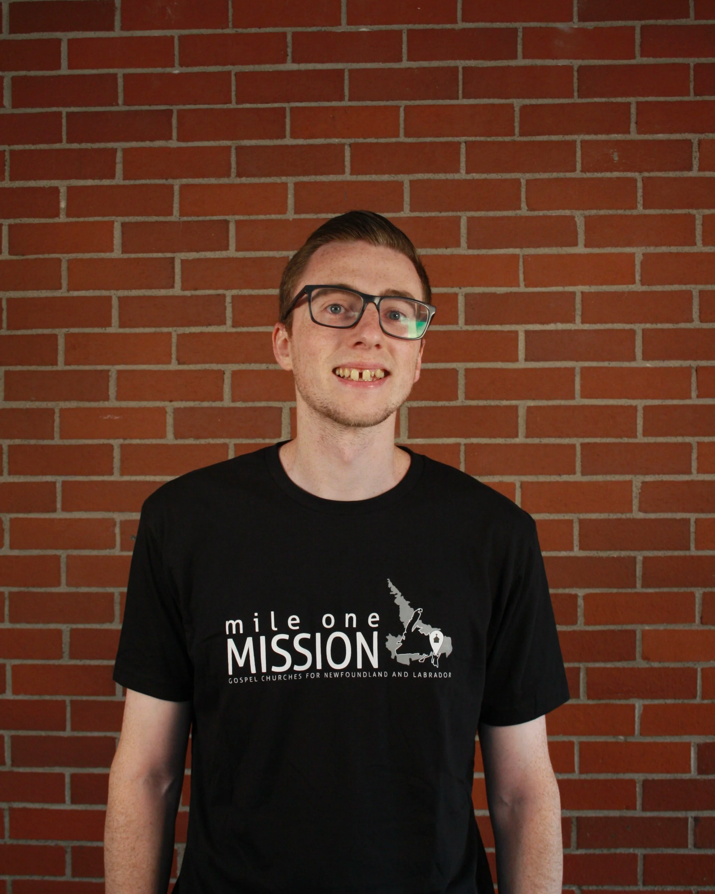 A young man with glasses smiling in front of a red brick wall, wearing a black t-shirt with the text 'mile one MISSION Gospel Churches for Newfoundland and Labrador' and a graphic of Newfoundland and Labrador.