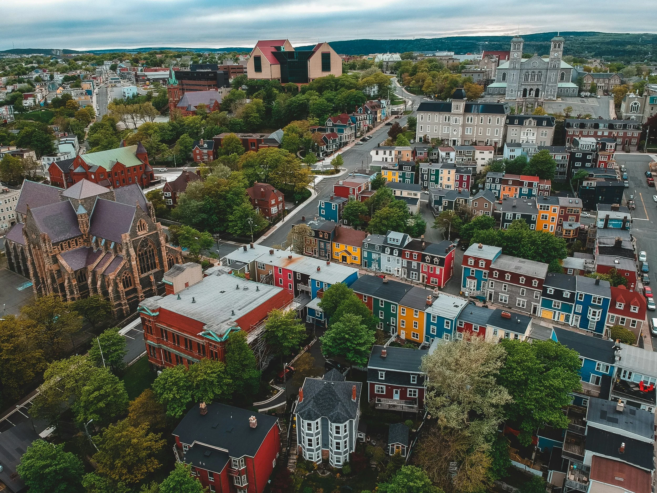 Aerial view of a small city with colorful houses, churches, and trees, with hills in the background under cloudy skies.