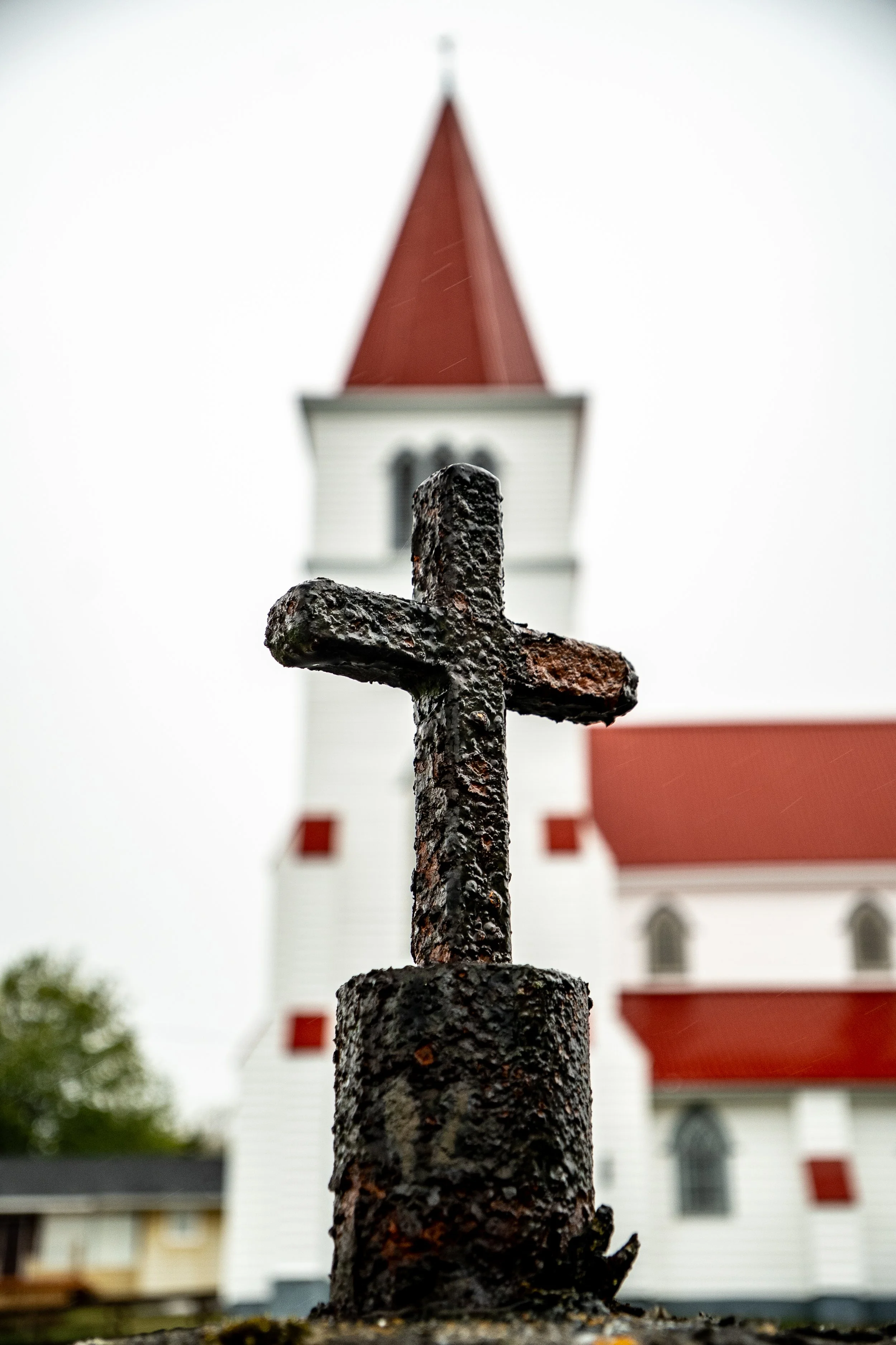 Close-up of a black, rusted iron cross on a church steeple with a white church with a red roof and a tall, tapered spire in the background.