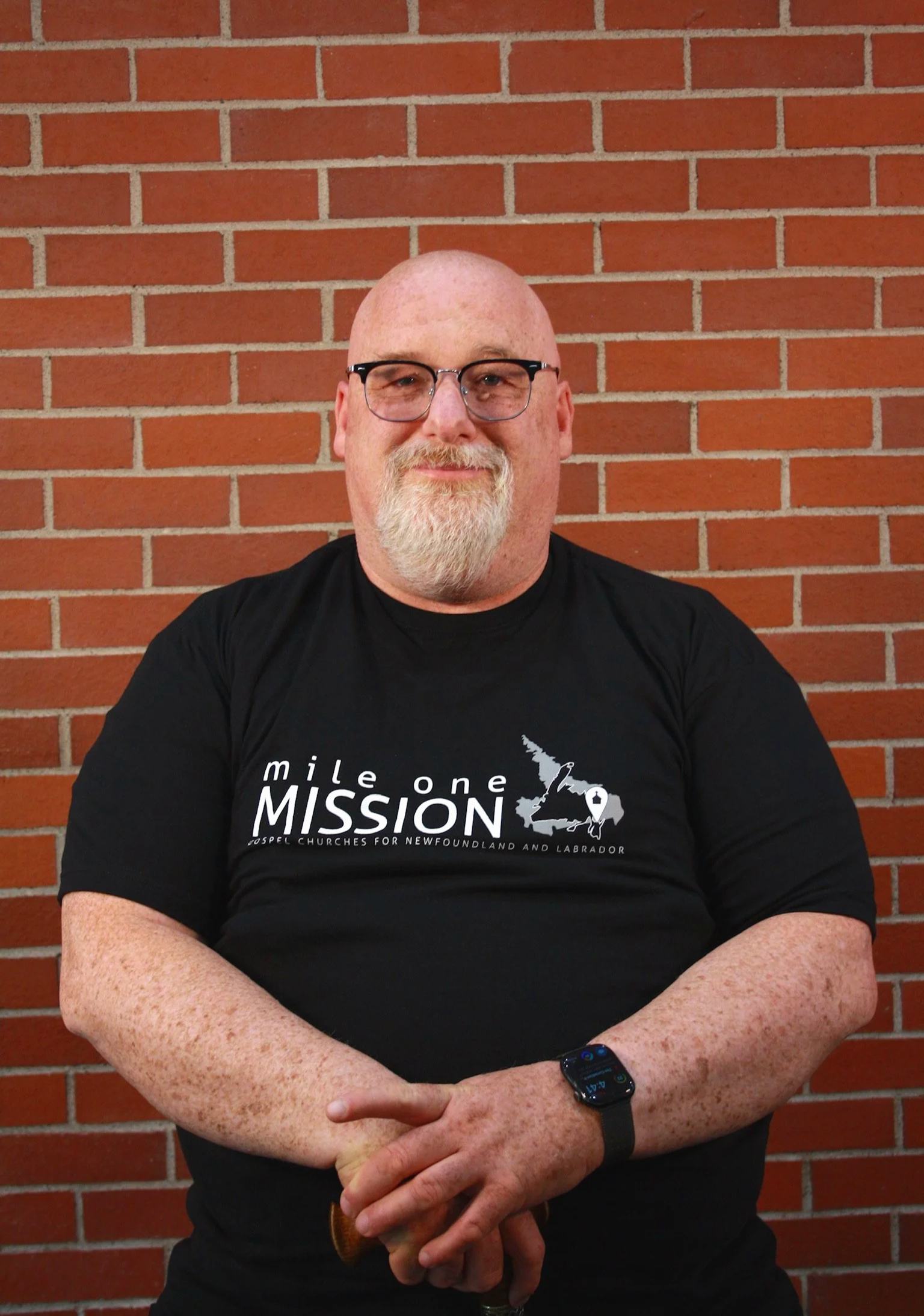 A bald man with glasses, a beard, and freckles is smiling and sitting against a red brick wall. He's wearing a black T-shirt and a smartwatch on his left wrist.