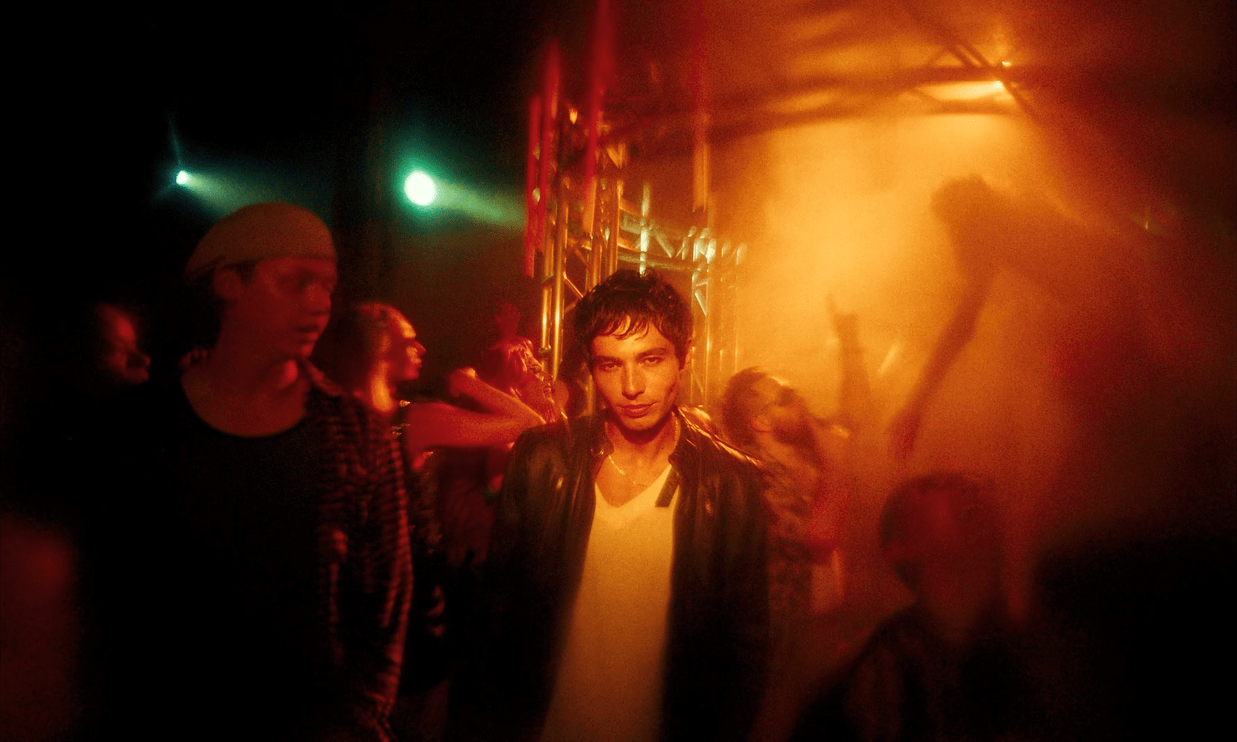 Young man with dark hair and leather jacket standing in a dimly lit nightclub with red and orange lighting, surrounded by other partygoers.