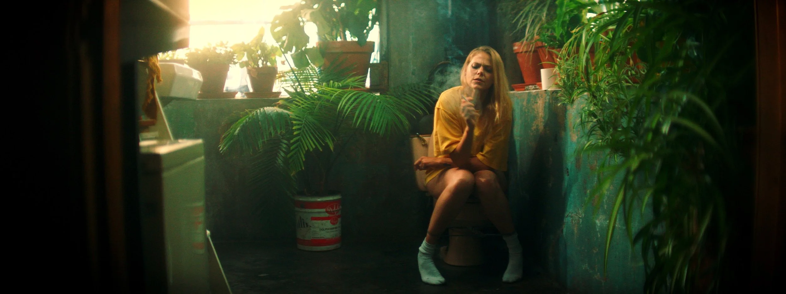 A woman in a yellow shirt sitting on a toilet in a cluttered, green, plant-filled bathroom, smoking a cigarette with sunlight streaming through a window.