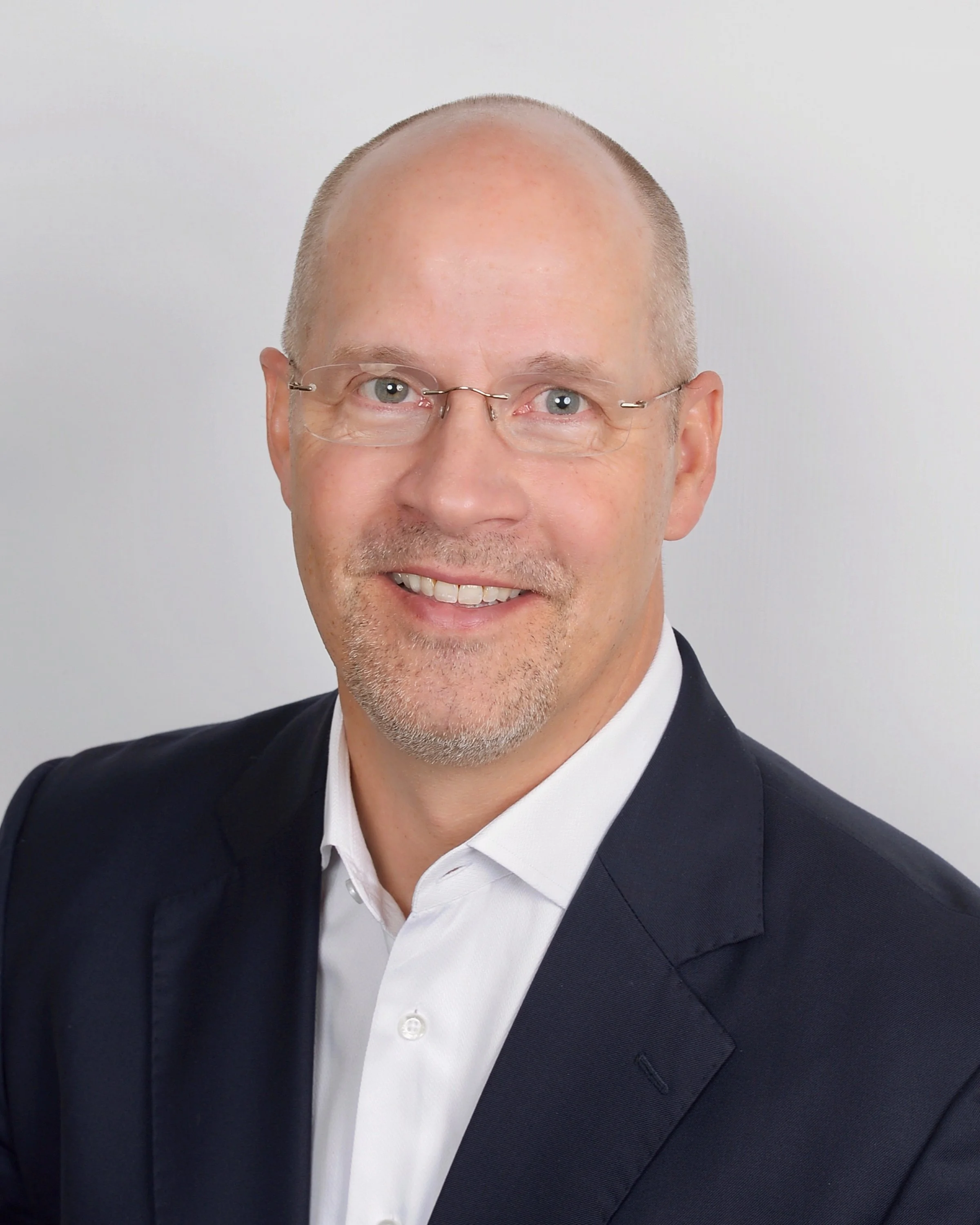 Close-up of a smiling man with short gray hair, wearing glasses, a dark suit, and a white shirt, against a plain light gray background.