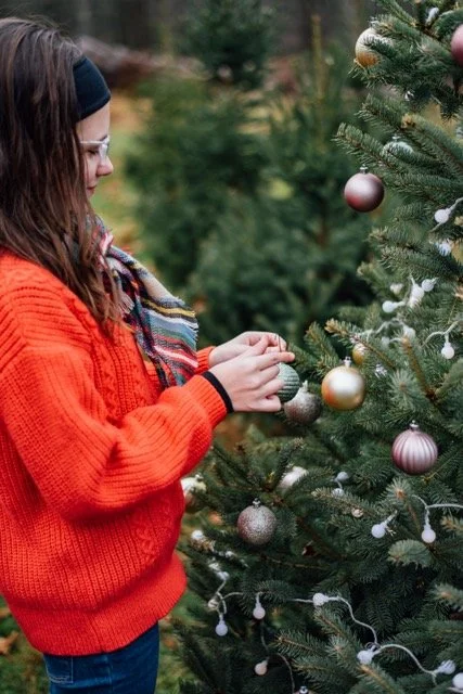 Girl in a red sweater and headband decorating a Christmas tree with ornaments outdoors.