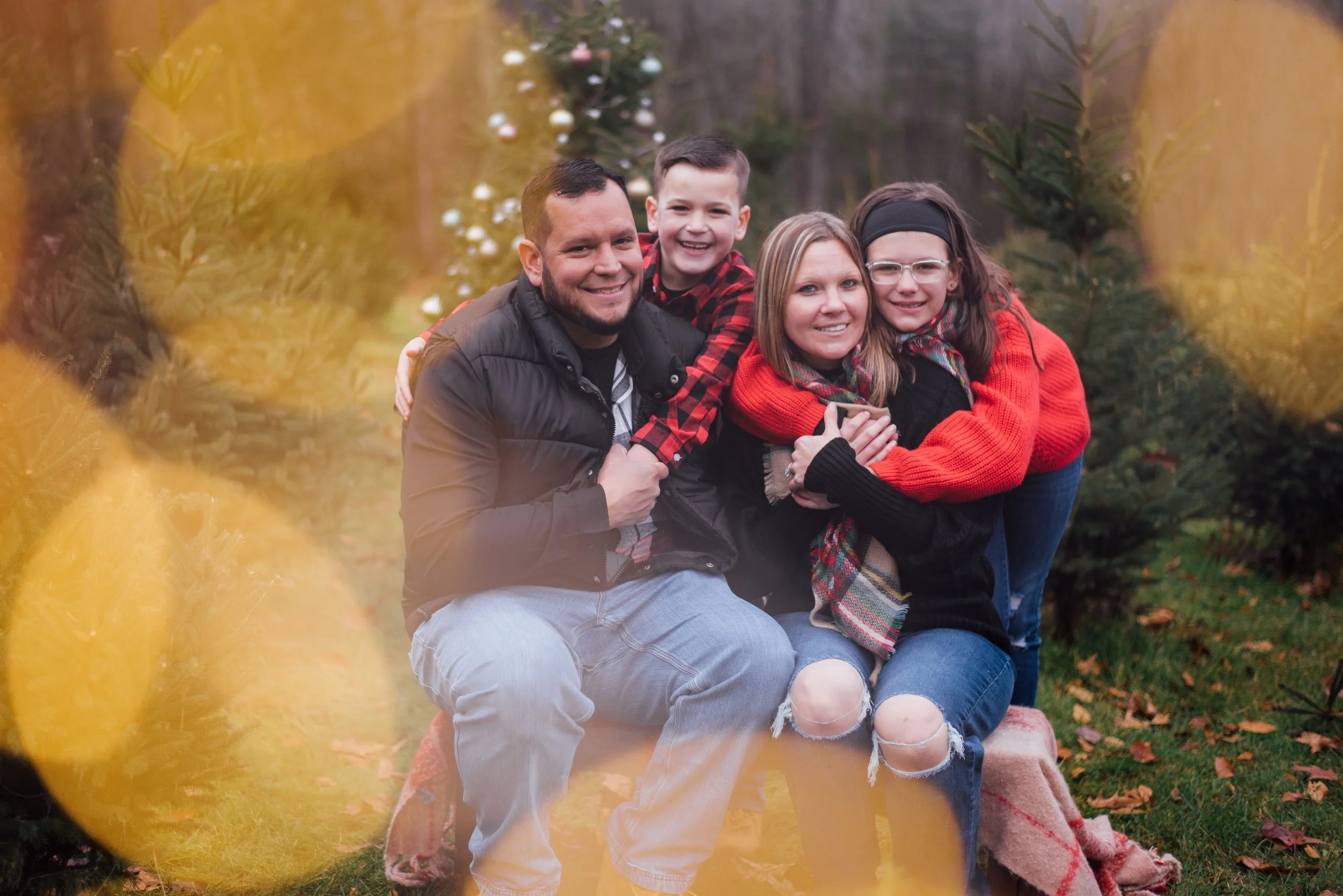 A family of four smiling and hugging outdoors during fall, with evergreen trees and a decorated Christmas tree in the background.