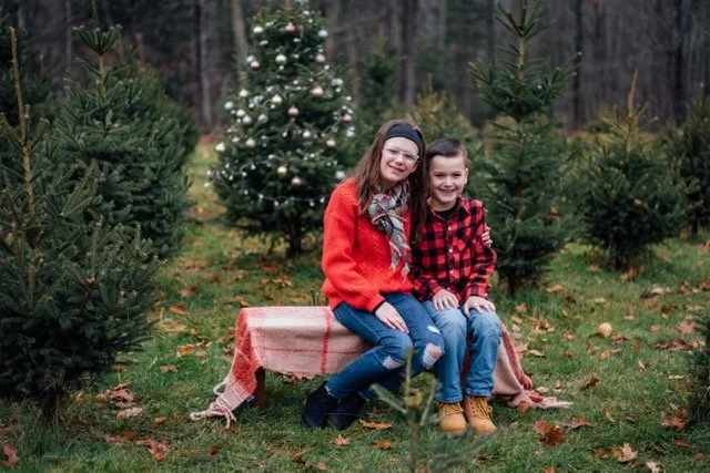 Two children, a girl and a boy, sitting on a bench outdoors in a Christmas tree farm with decorated and unadorned evergreen trees, smiling and dressed in festive red and black plaid clothing.