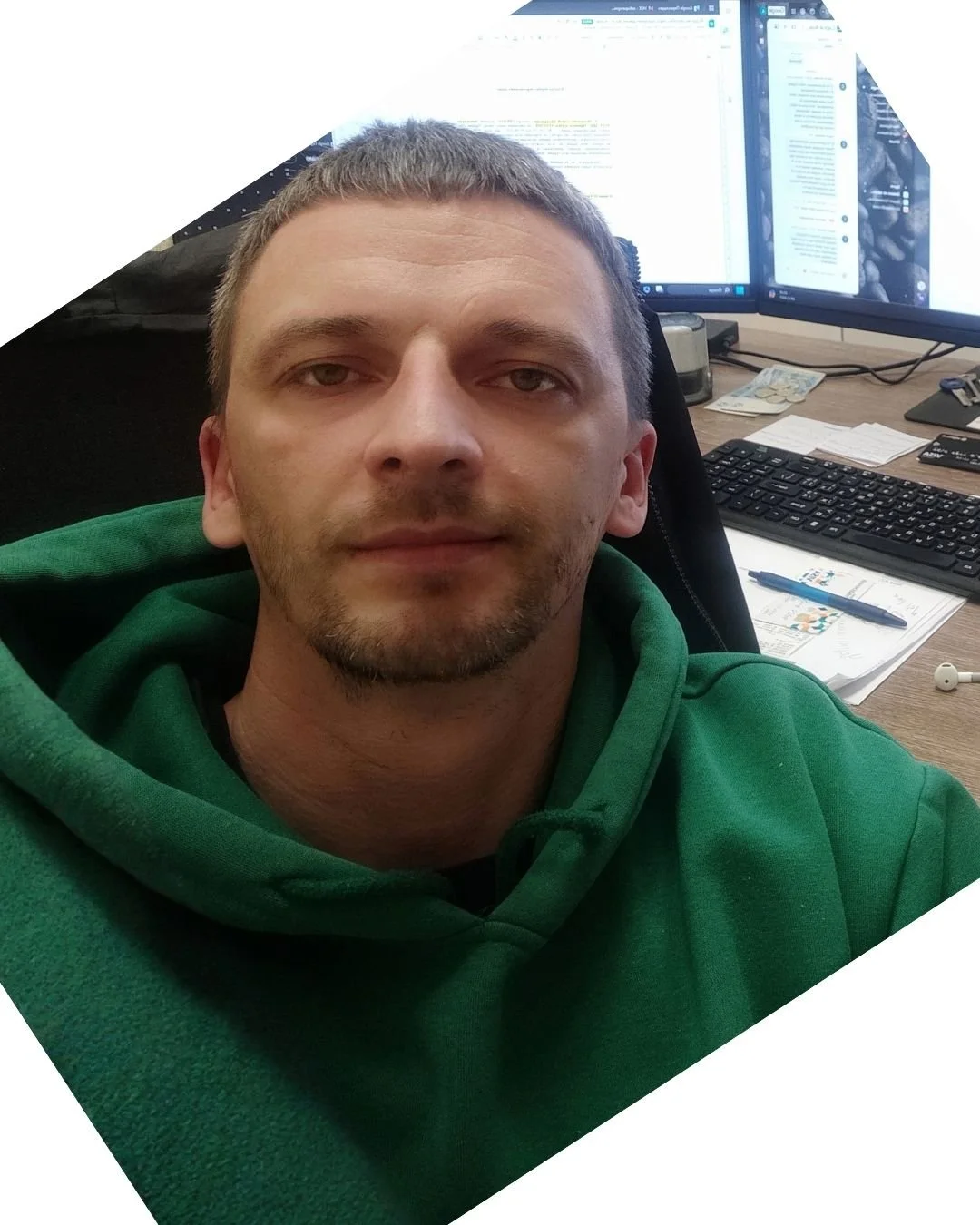A man wearing a green hoodie sitting at a desk with two monitors, a keyboard, papers, a pen, and earphones.