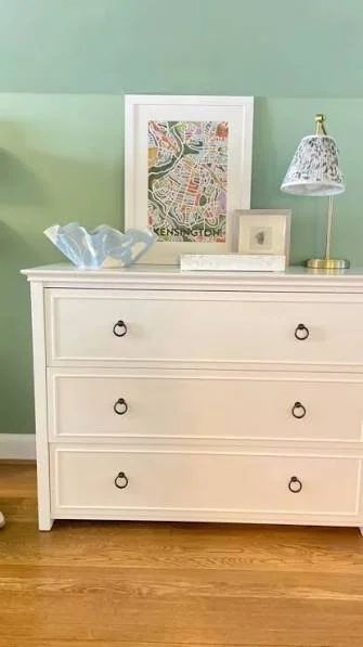 A white dresser with four drawers, each with two round metal handles, is positioned against a light green wall. On top of the dresser, there is a framed artwork, a small picture frame, a lamp with a patterned shade, and a decorative glass bowl.