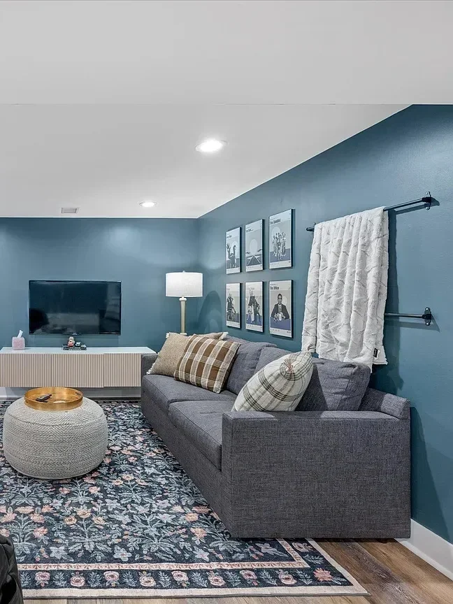 Living room with a gray sofa, a plaid throw pillow, a beige ottoman with a tray, a blue wall with framed pictures, a white curtain, a floor lamp, a television, and a patterned area rug.