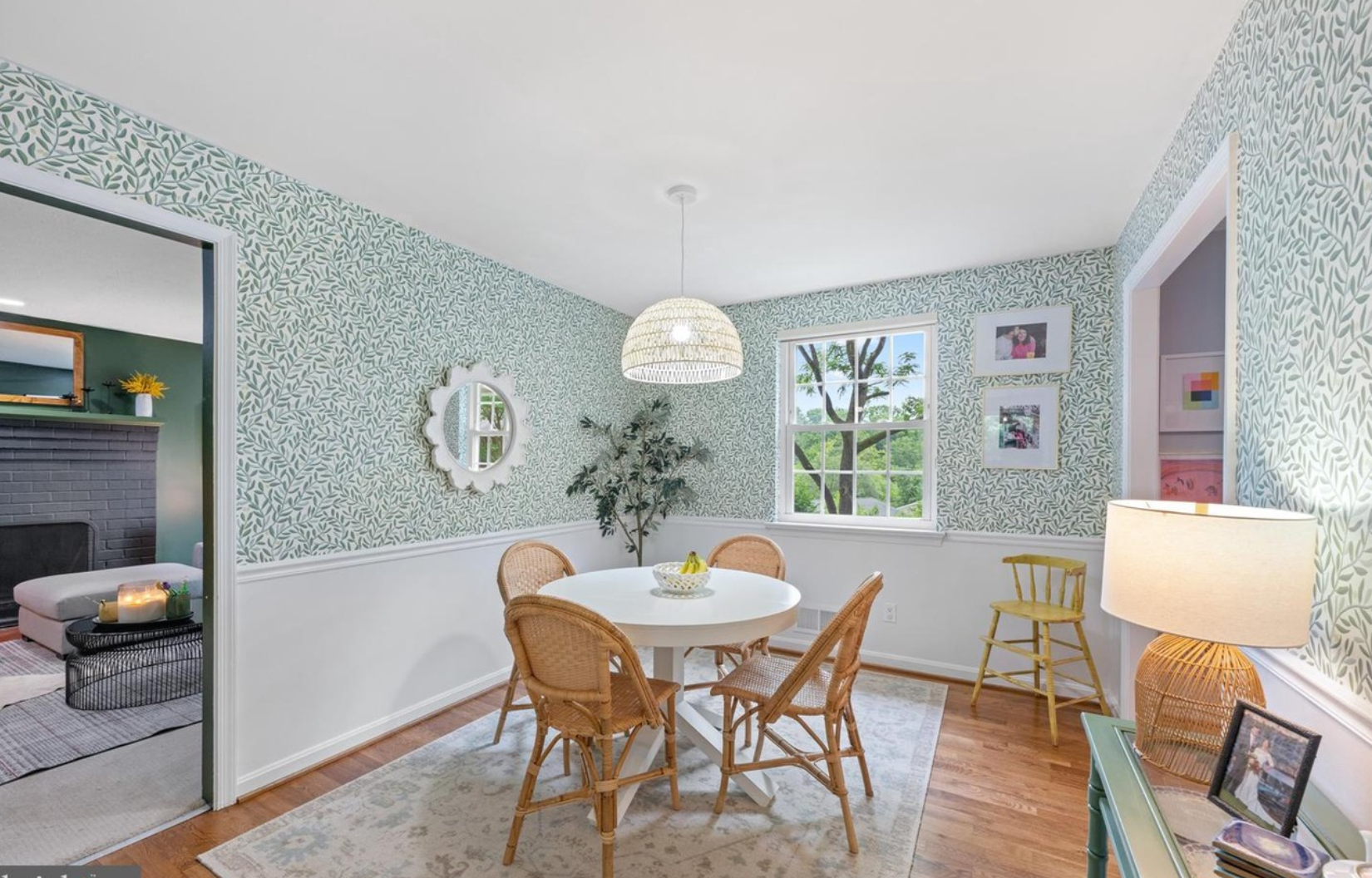 A dining room with a round white table and four wicker chairs. Light green leafy wallpaper, a white ornate mirror on the wall, a window showing trees outside, and a potted plant. A rug, a yellow chair, and a lamp are also visible.