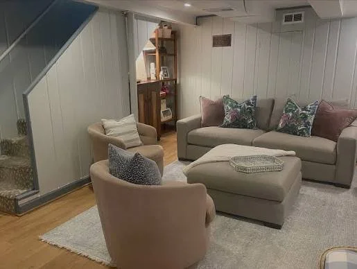 Living room with beige armchair, gray sofa with floral pillows, ottoman, and bookshelf.
