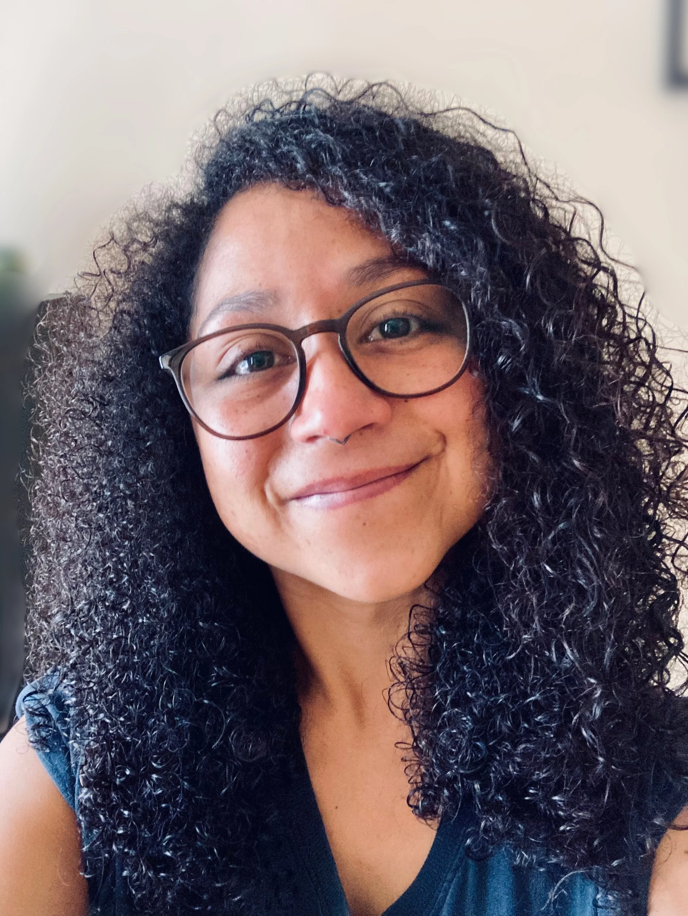 Close-up of a woman with curly dark hair, glasses, and a warm smile, wearing a blue top.