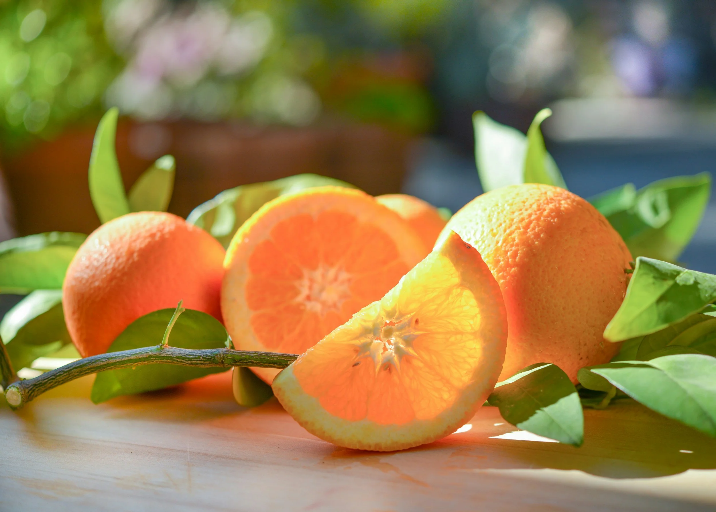 Close-up of fresh oranges, some whole and some sliced, with green leaves and a branch, on a light wooden surface with a blurred outdoor background.