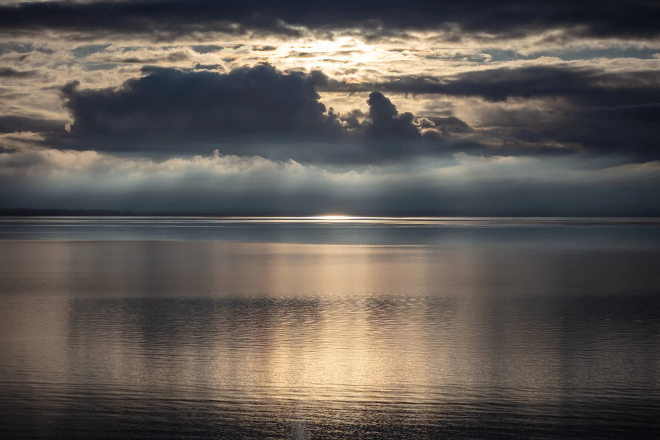 Et hav med overskyet himmel hvor sollys bryder igennem skyerne over vandet. Kalø Vig