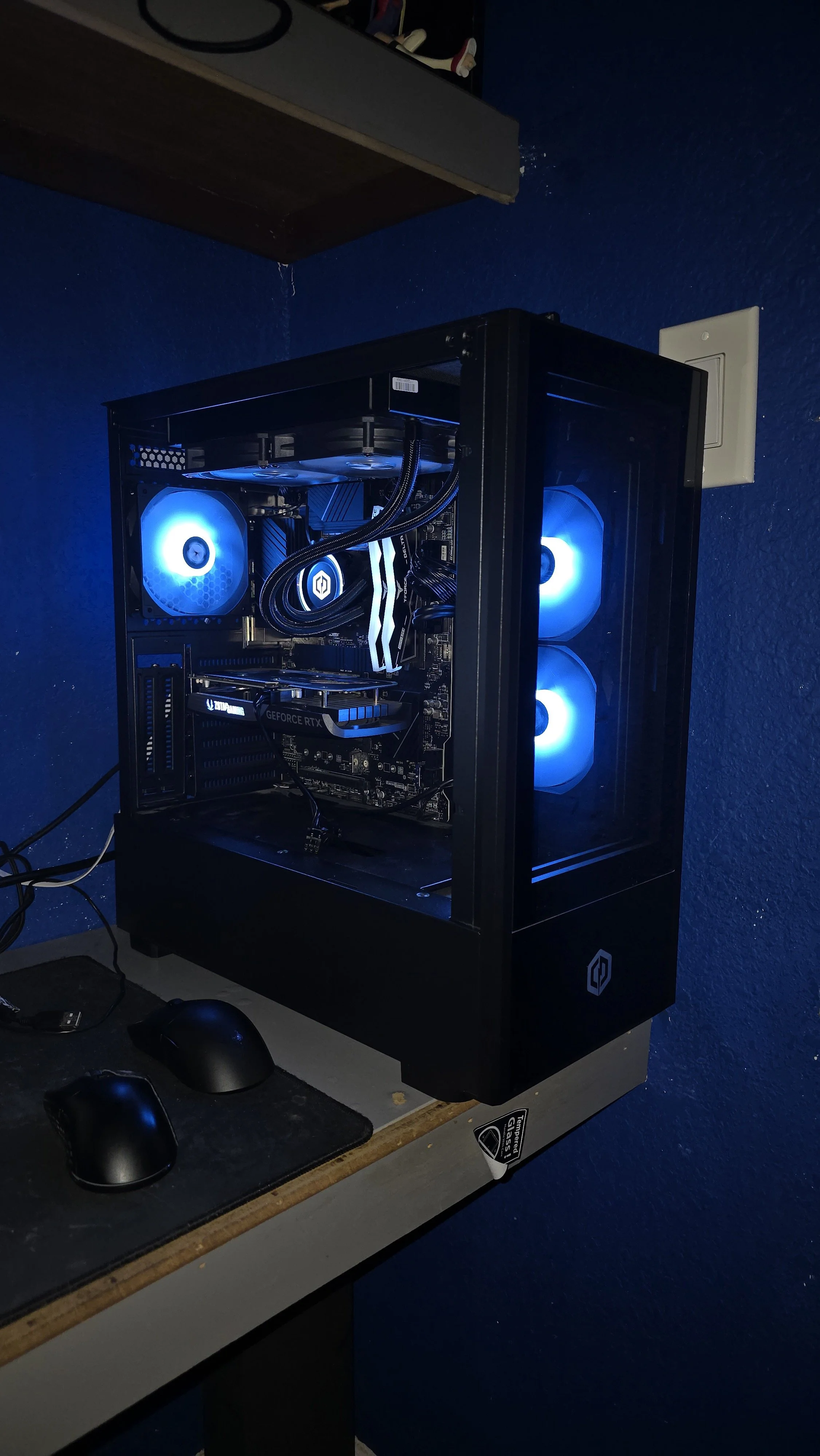 A gaming or high-performance computer tower with blue LED lights inside, placed on a desk with a mouse and mouse pad, against a blue wall and a wooden shelf above.