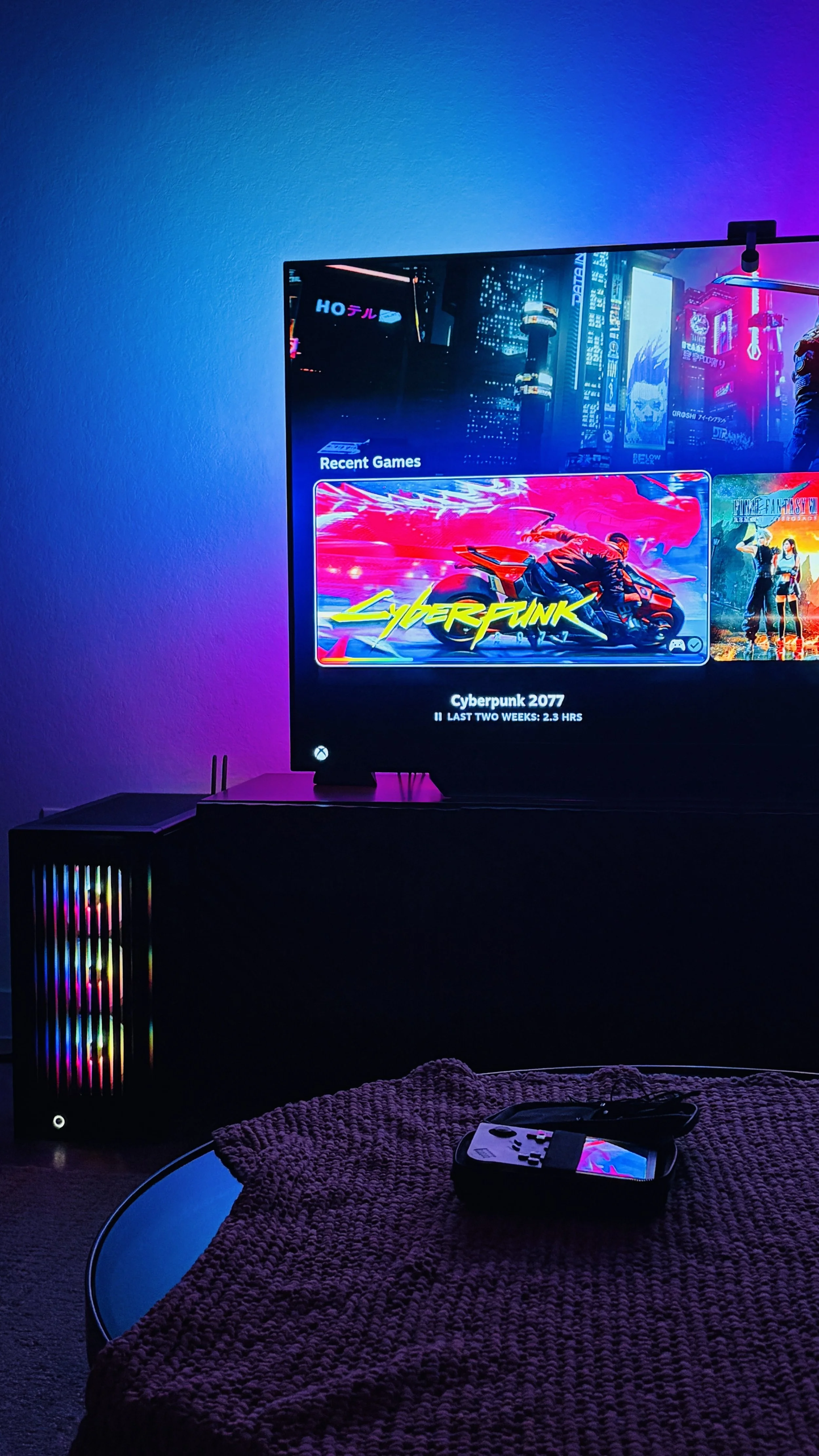 A gaming setup with a large flat-screen TV displaying a screen with the game Cyberpunk 2077, surrounded by colorful LED lighting. There is a gaming console or PC beneath the TV with a colorful LED-lit tower. A game controller and a TV remote are on a round table covered with a pink textured cloth in the foreground.
