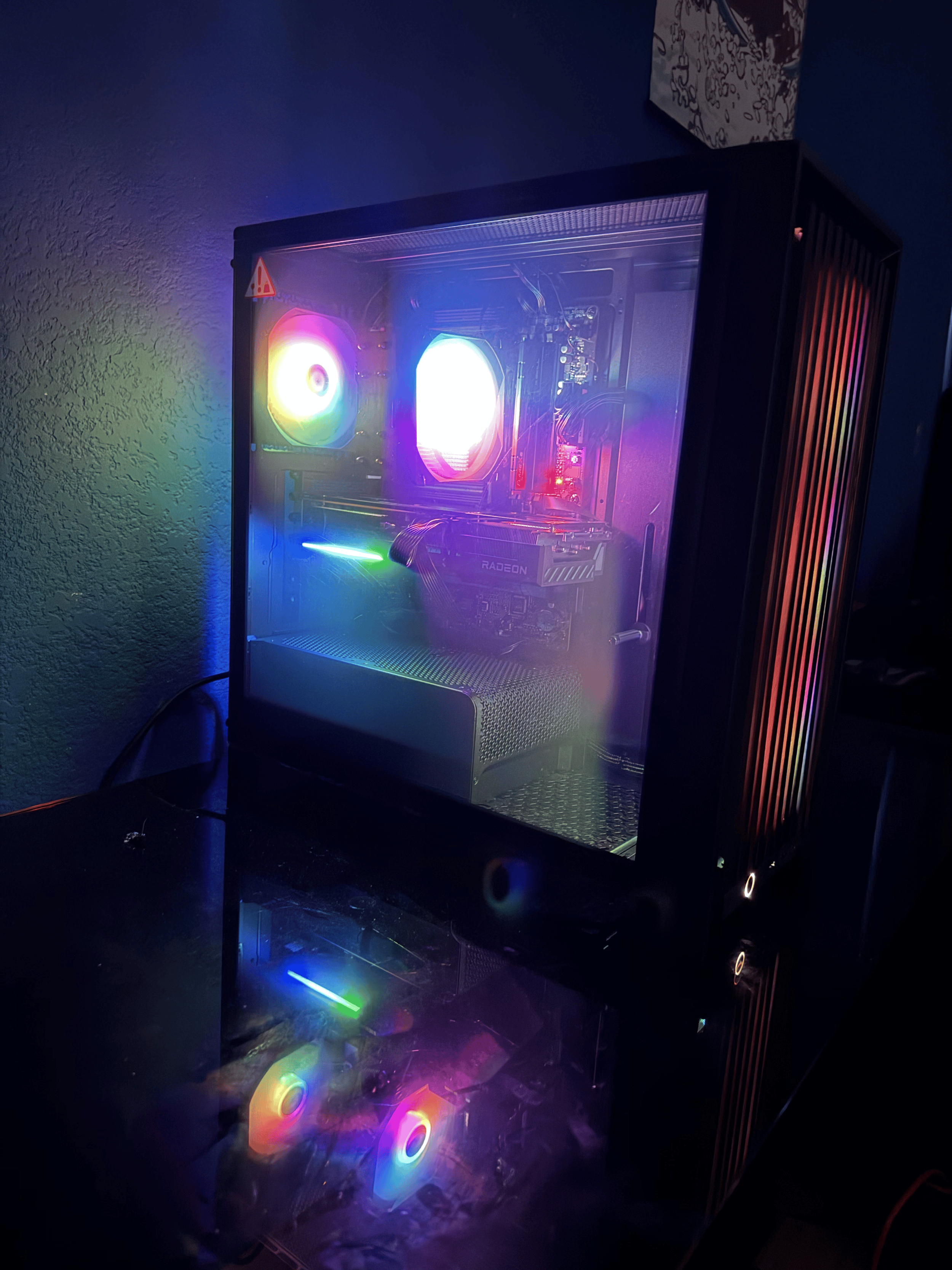 A gaming computer with internal RGB lighting visible through a transparent side panel. The case features colorful LED fans and a graphics card with the brand 'Radeon' visible, set against a dark room background.