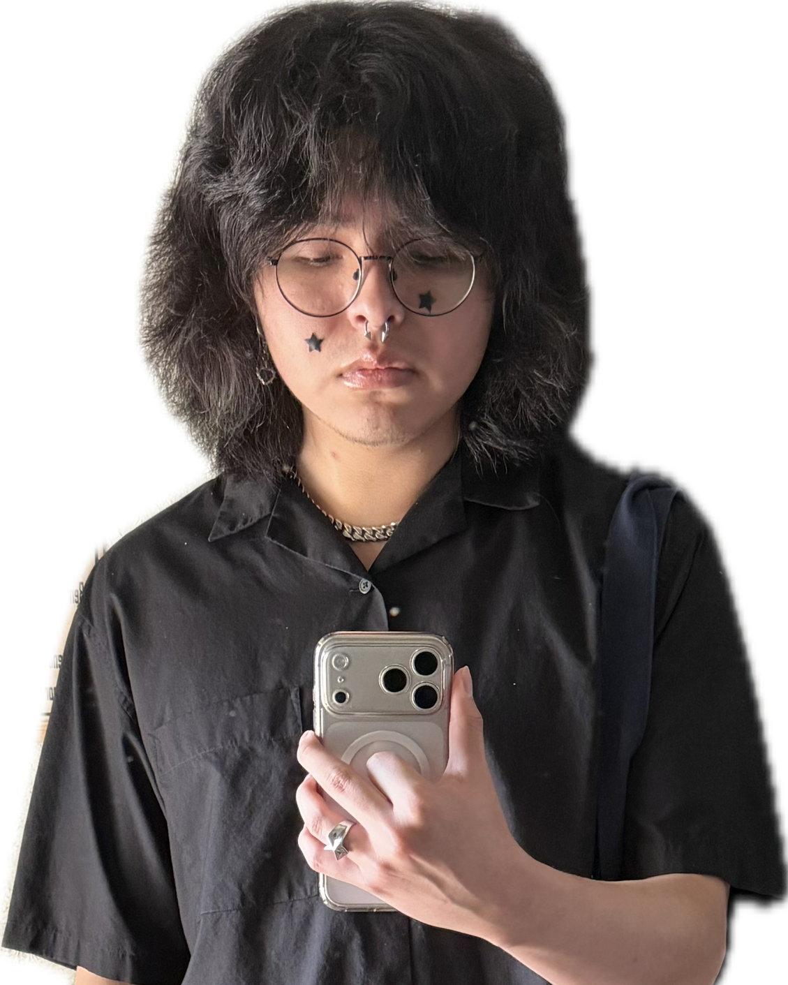 A person with shoulder-length, wavy dark hair, wearing glasses, star-shaped facial tattoos, and silver jewelry, taking a selfie with a smartphone.