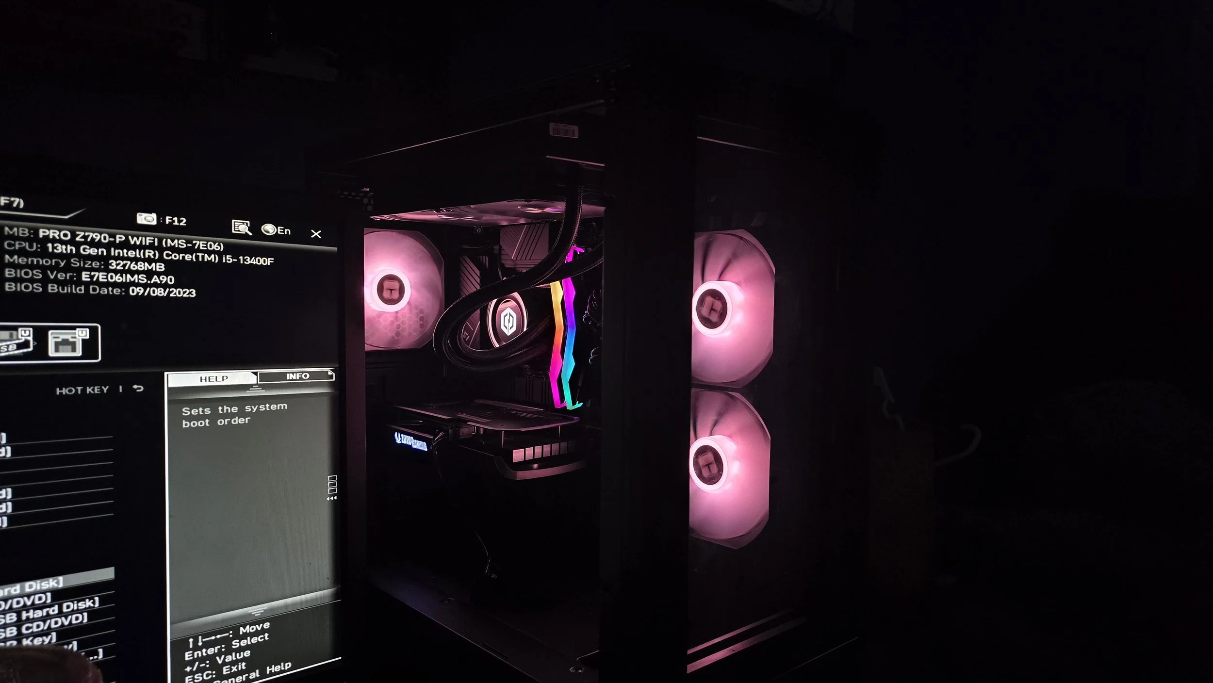 A computer setup with internal components visible, including pink illuminated cooling fans and RGB lighting on the motherboard. A monitor displays system information and BIOS settings.