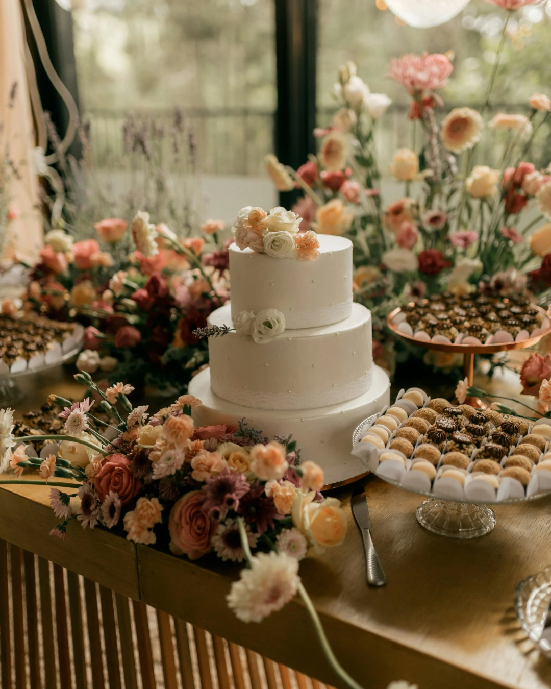 A three-tiered white wedding cake with floral decorations on top, displayed on a wooden table surrounded by assorted flowers and desserts at a celebration.