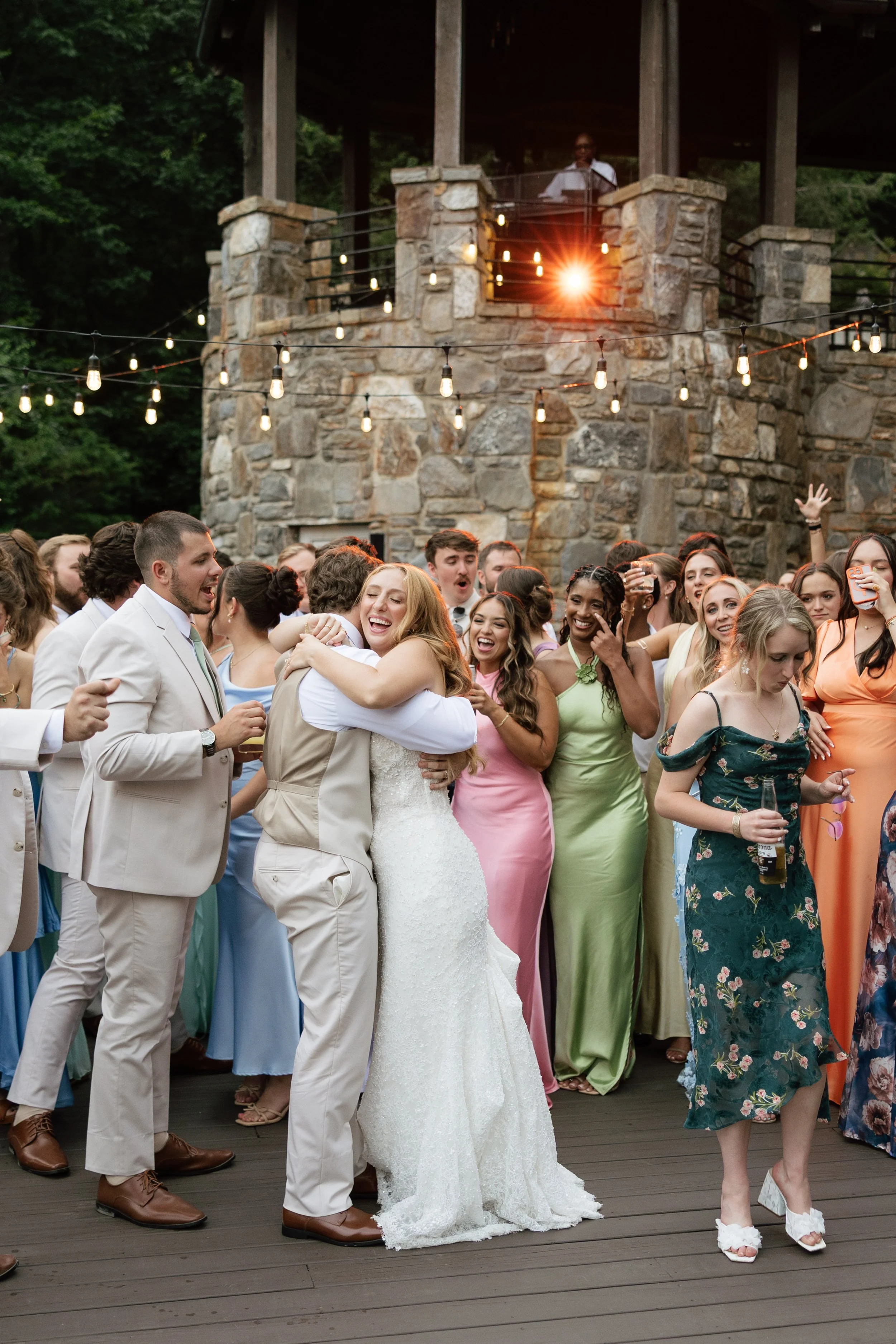 A large group of people dancing and celebrating outdoors on a wooden deck at a wedding reception, with string lights overhead and a stone building in the background. The bride and groom are at the center, hugging each other and smiling.