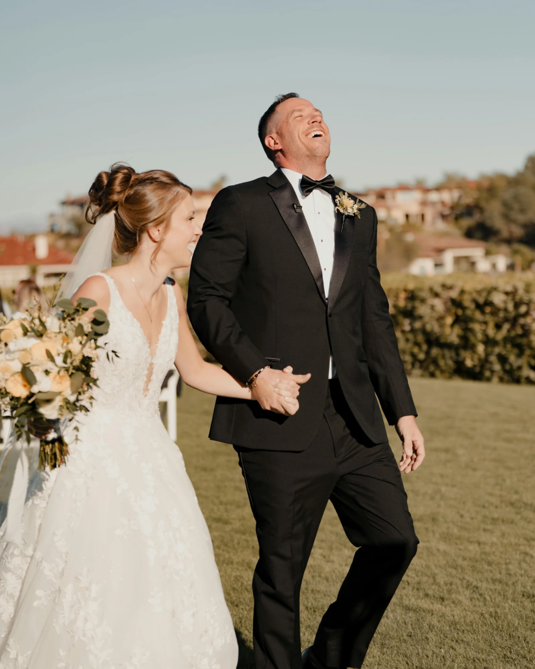 A bride and groom holding hands and laughing outdoors on their wedding day.