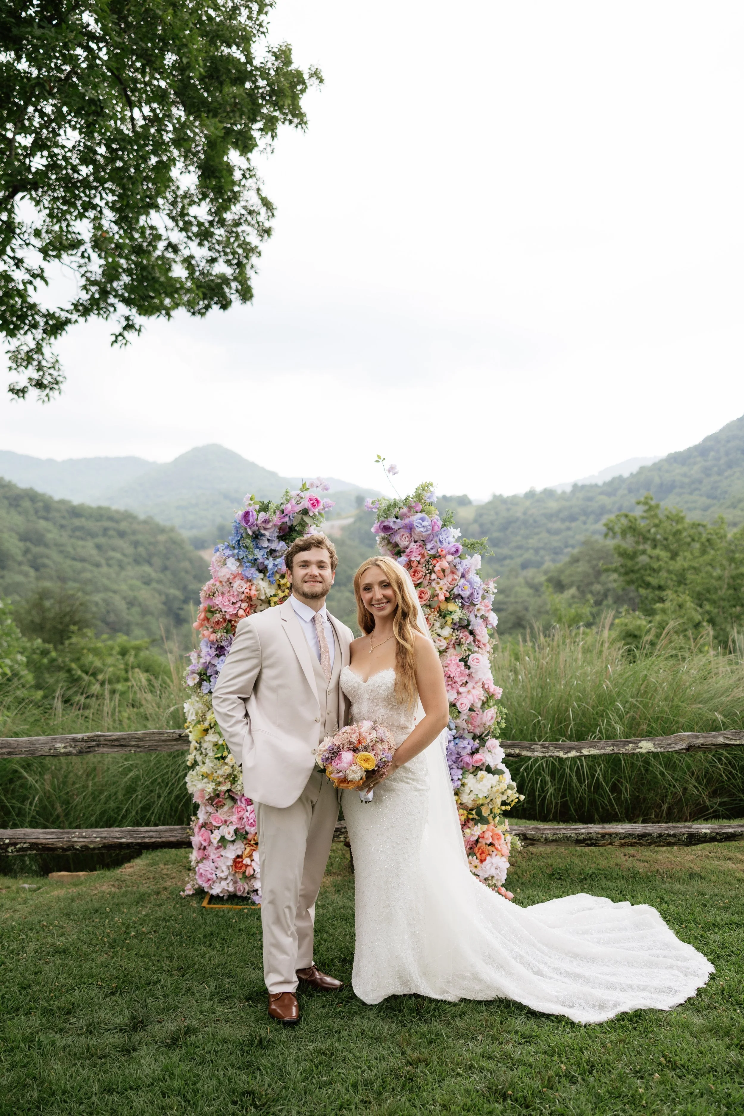 A newlywed couple standing outdoors in front of a floral arch, with mountains in the background. The groom is wearing a beige suit and tie, while the bride is in a white lace wedding gown holding a bouquet. They are smiling, and the scenery includes green trees and grass.