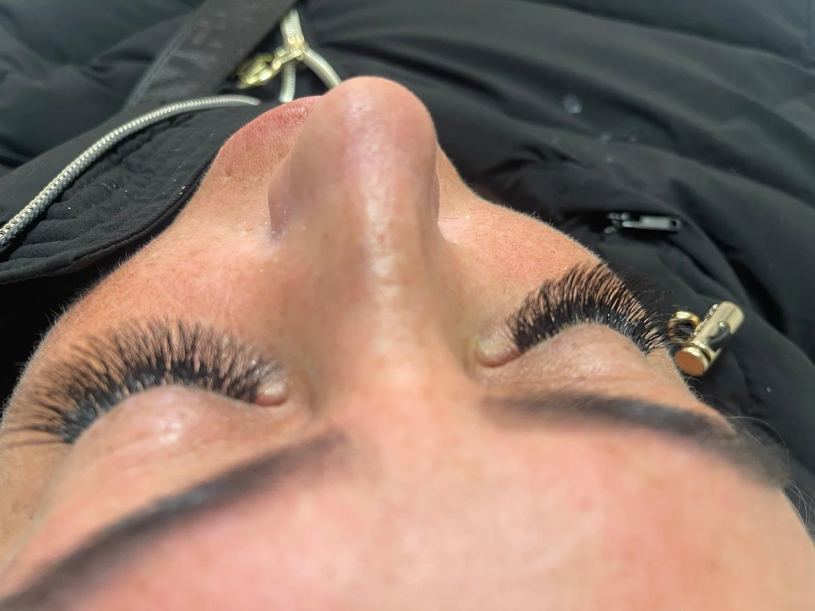 Close-up of a woman's face with long, thick eyelashes, lying down with her eyes closed, wearing black clothing with gold zipper details.