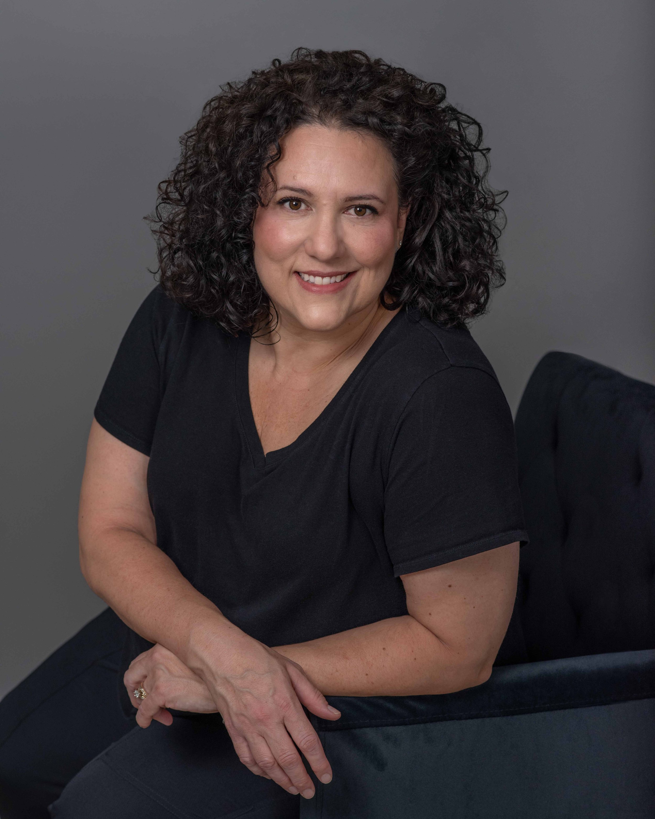 A woman with shoulder-length curly dark hair, wearing a black shirt, smiling, seated with her arms crossed on a dark surface against a gray background.