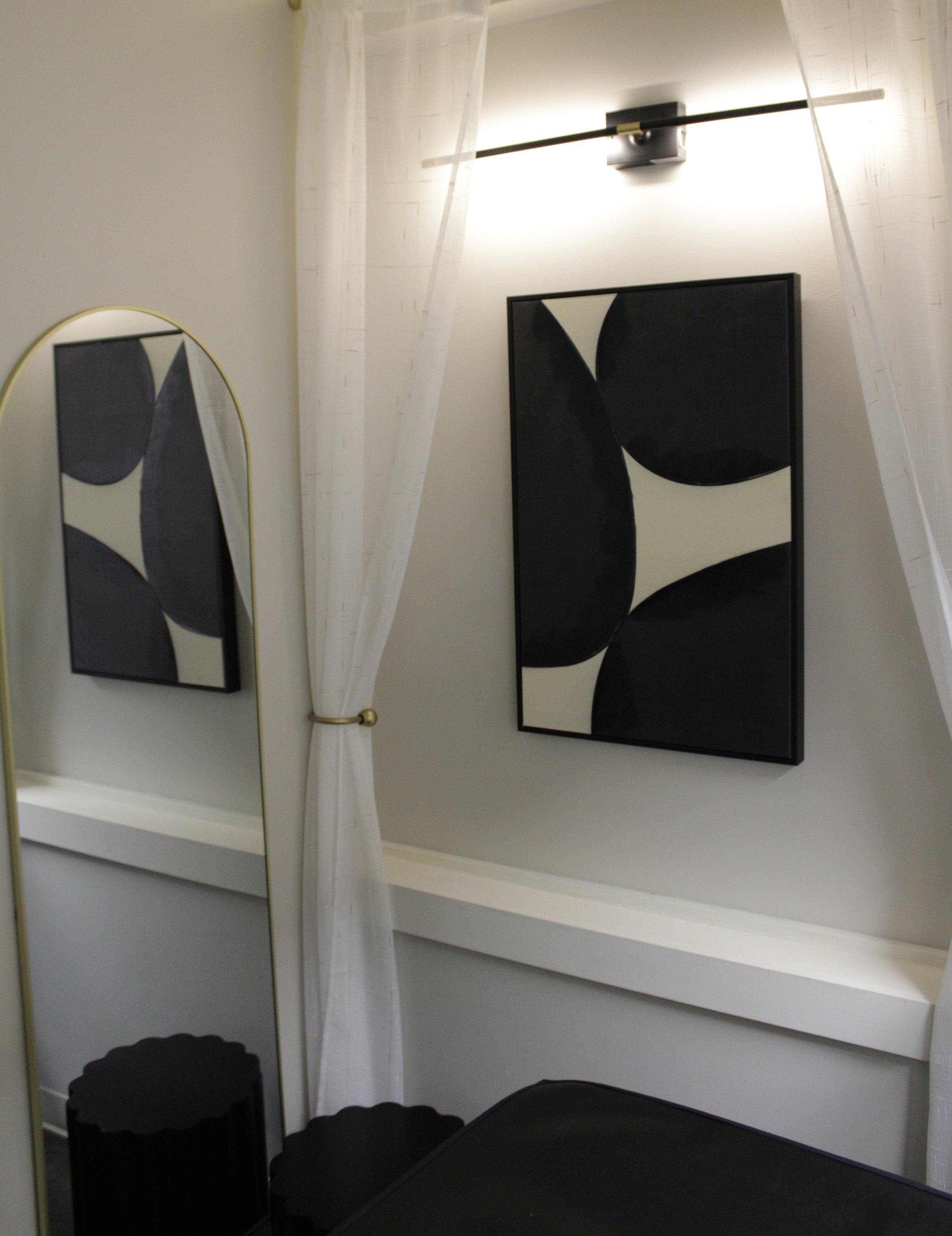 Interior of a minimalist room with a black and white abstract painting on a wall, a curved mirror with a gold frame, sheer white curtains, and black furniture.