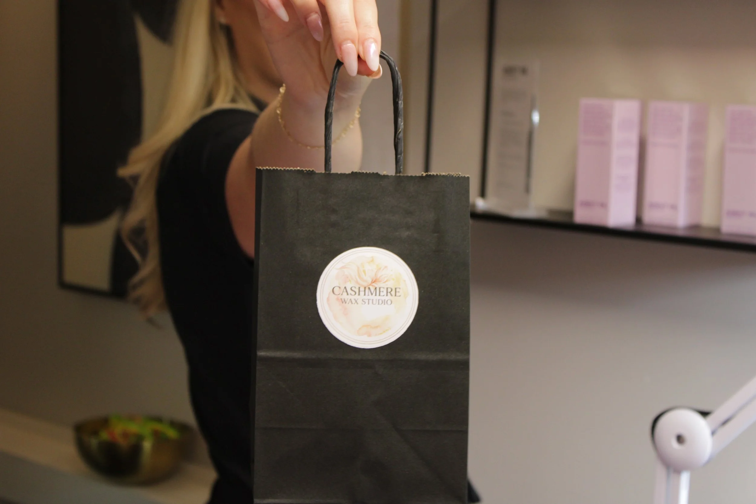 A person holding a black shopping bag with a round sticker that reads 'Cashmere Wax Studio,' in a retail or skincare setting.