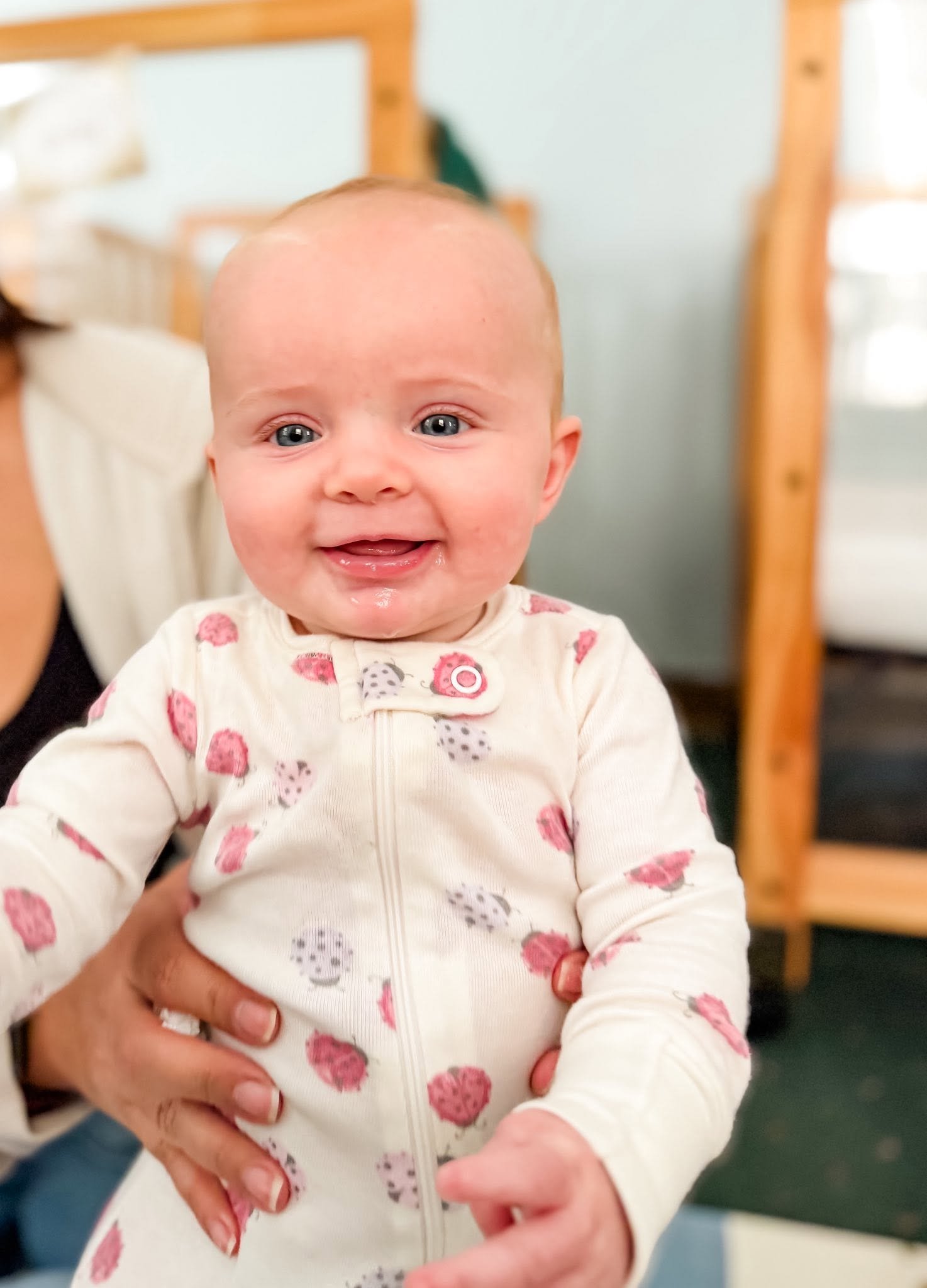 Close-up of a smiling baby wearing a cream onesie with pink and gray berry patterns, being held by an adult in an indoor setting.